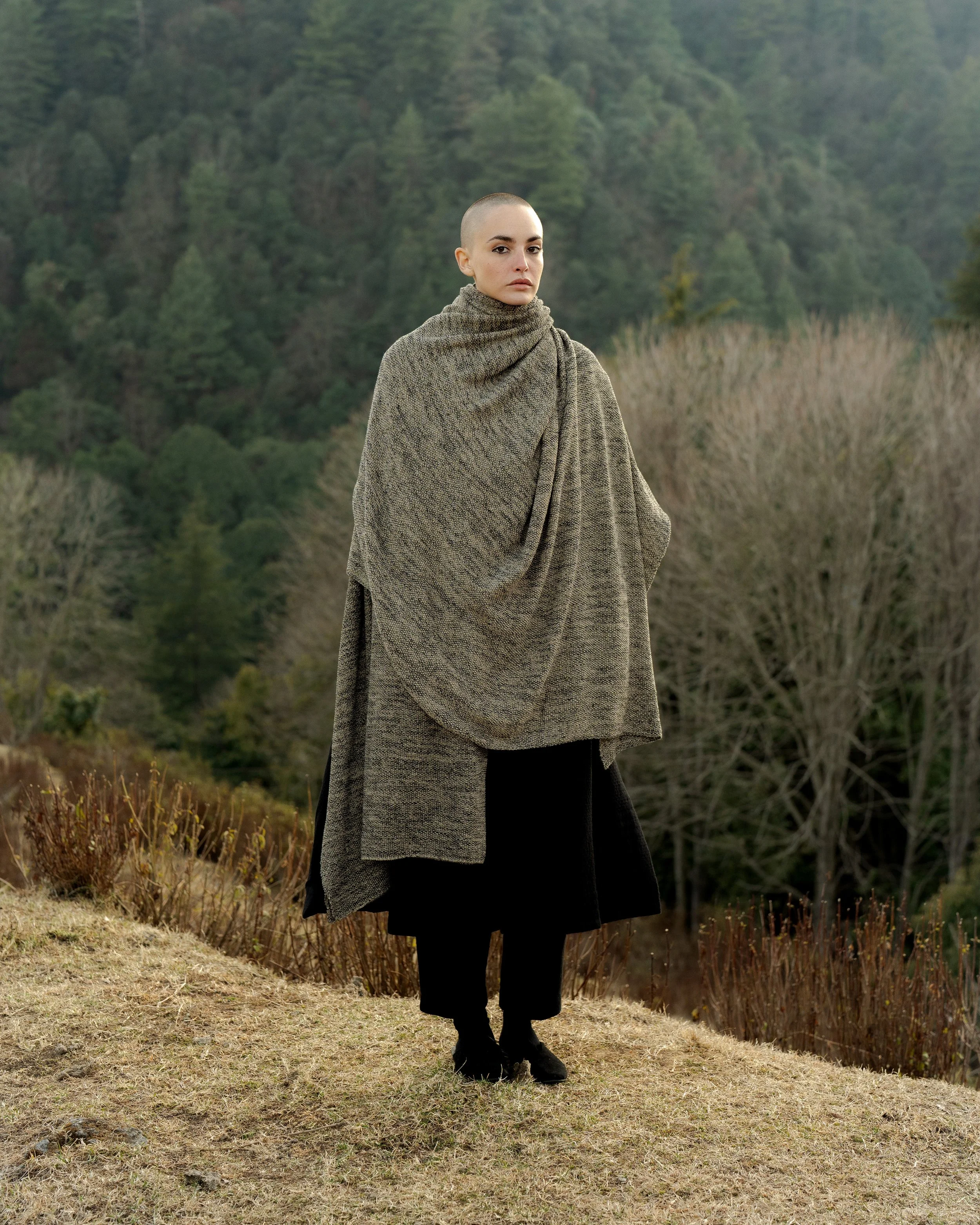 A person with a shaved head and light makeup stands outdoors in a field, wearing a large, textured, taupe-colored shawl and black shoes, with a forested hillside in the background.