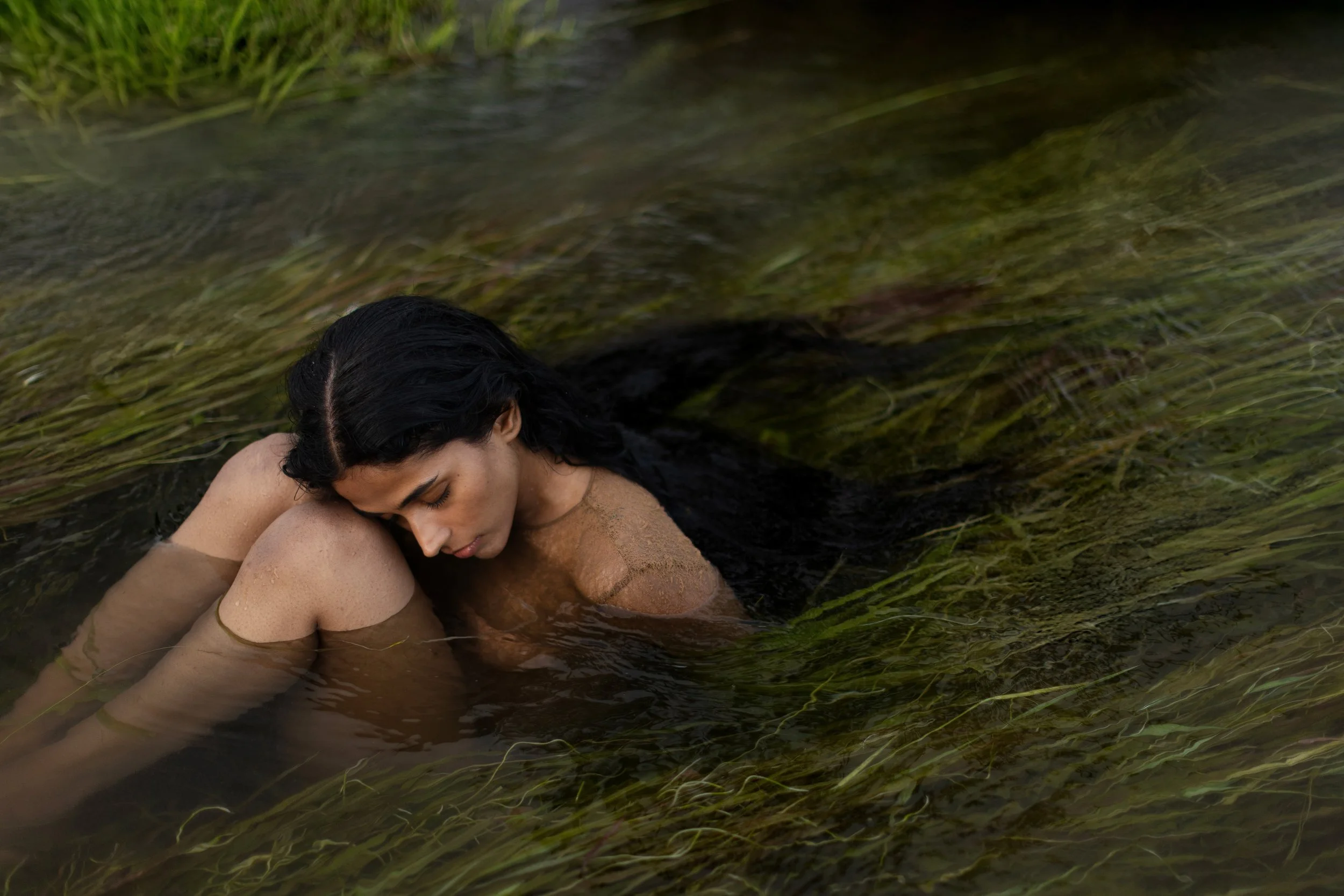 Woman with dark hair submerged in water, leaning forward with her eyes closed, surrounded by underwater grass and reeds.