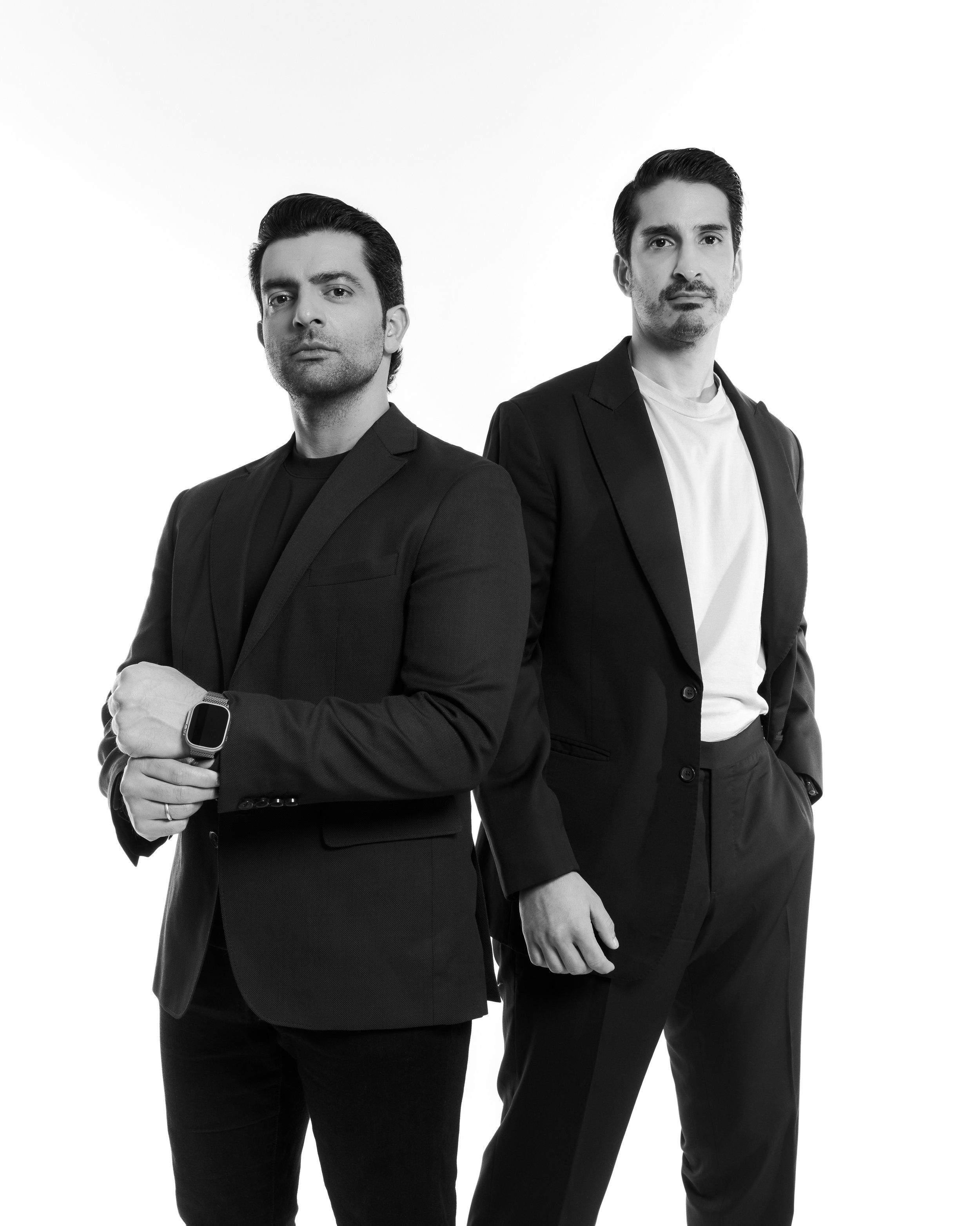 Two men in business attire stand confidently against a plain white background, with one man in the foreground and the other in the background.