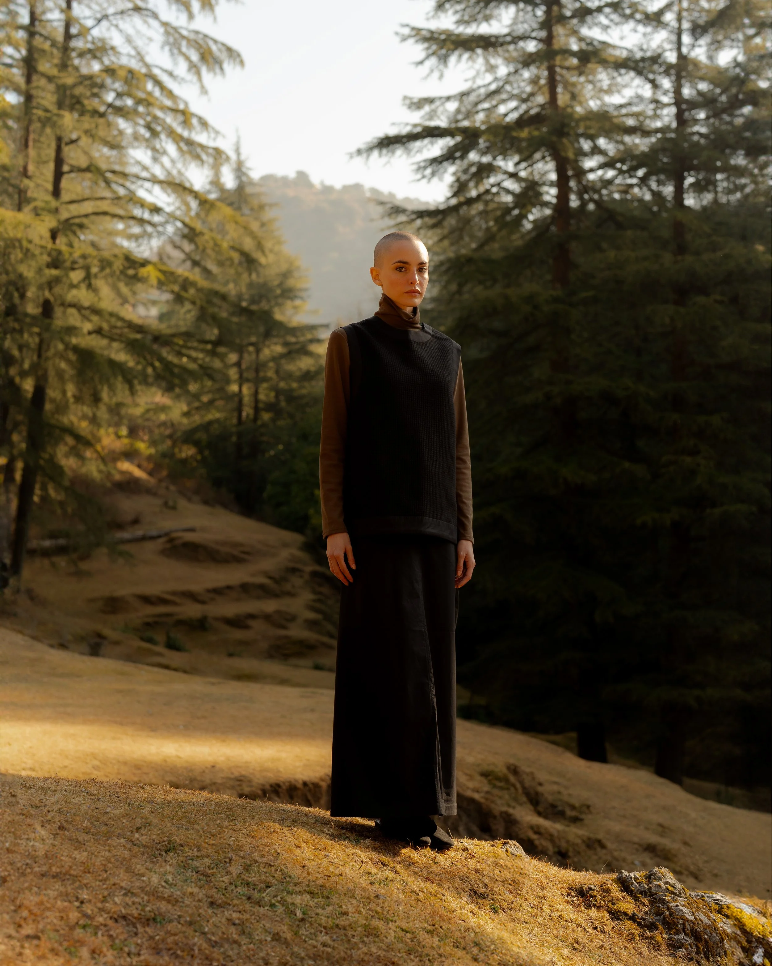 A woman with a shaved head wearing a black top and brown sleeves standing outdoors on a hillside with trees in the background during daytime.