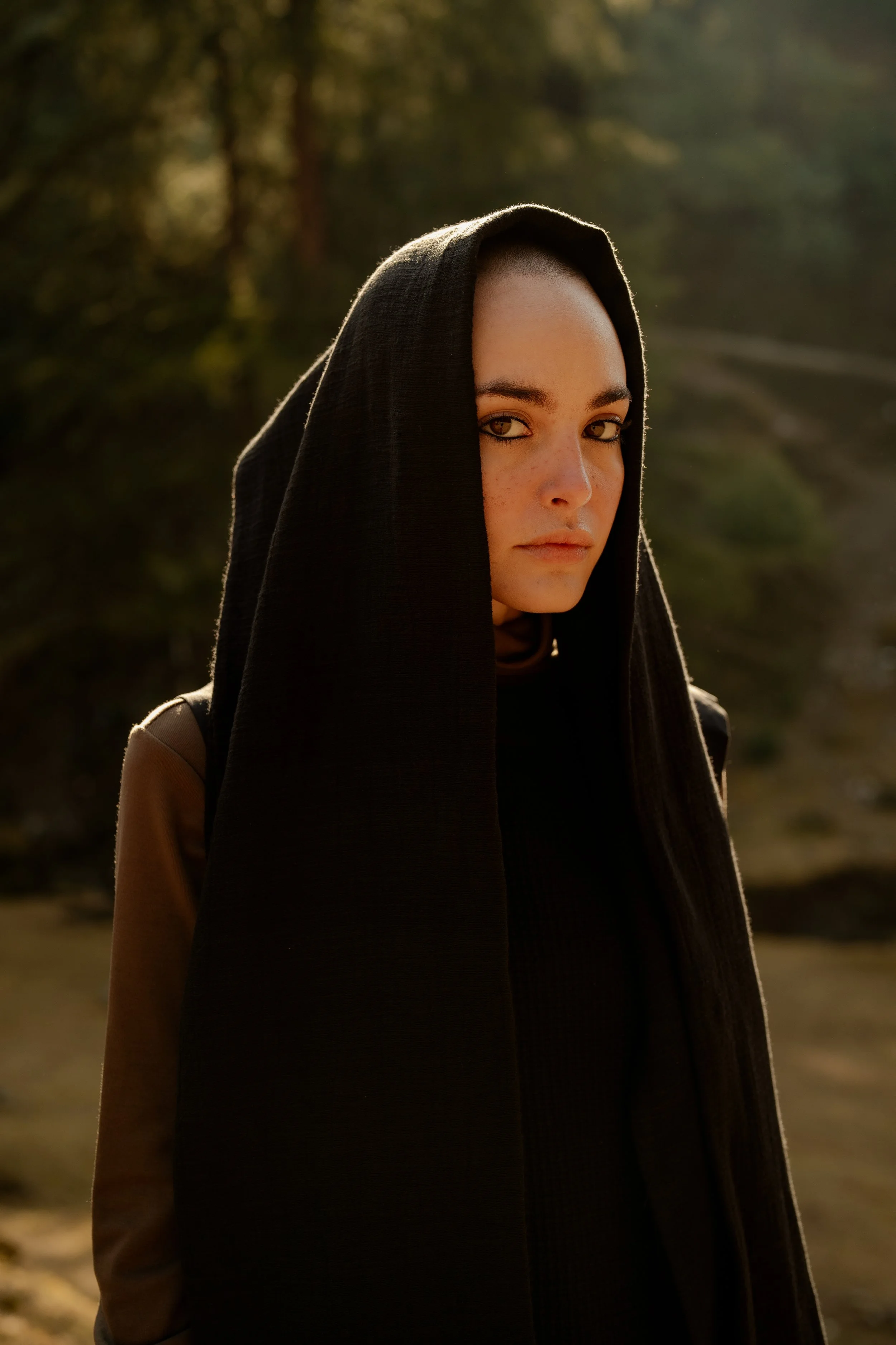 A woman with dark hair and light skin wearing a black hooded garment in an outdoor setting during golden hour.