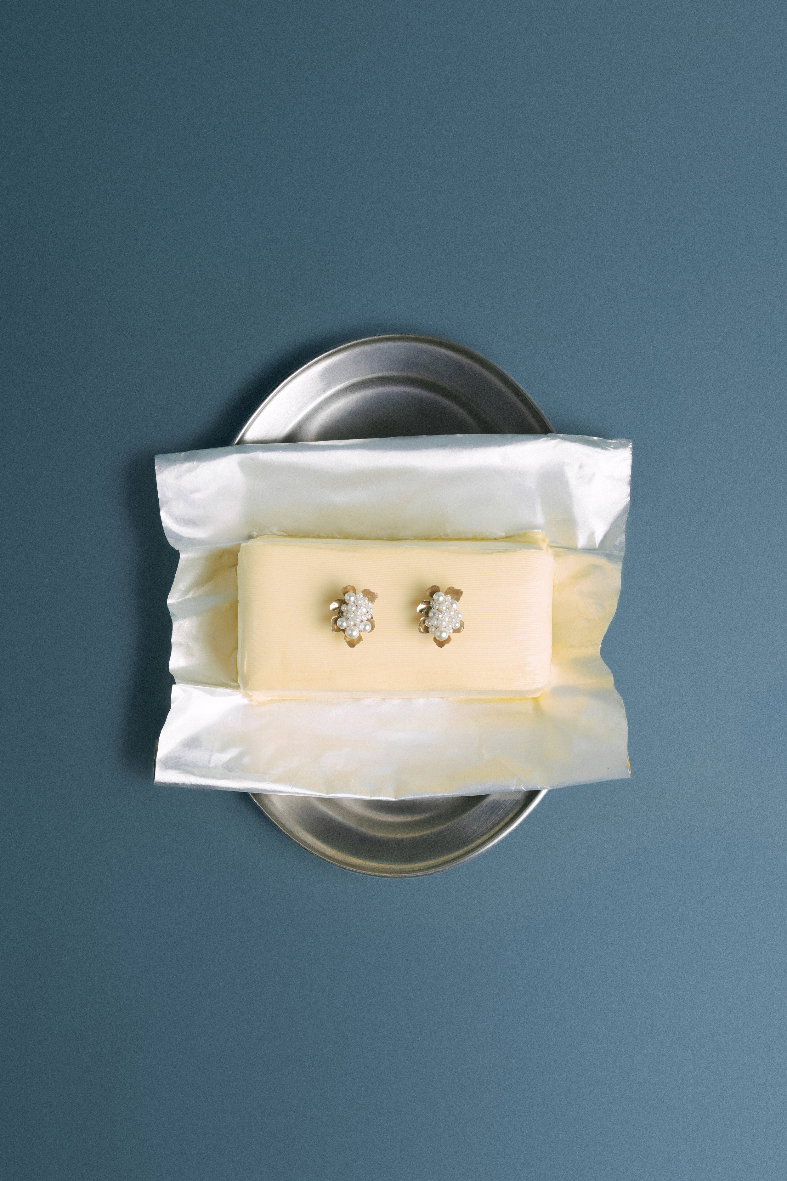 A pair of pearl earrings on a white cushion, partially covered by a metallic container, against a gray background.