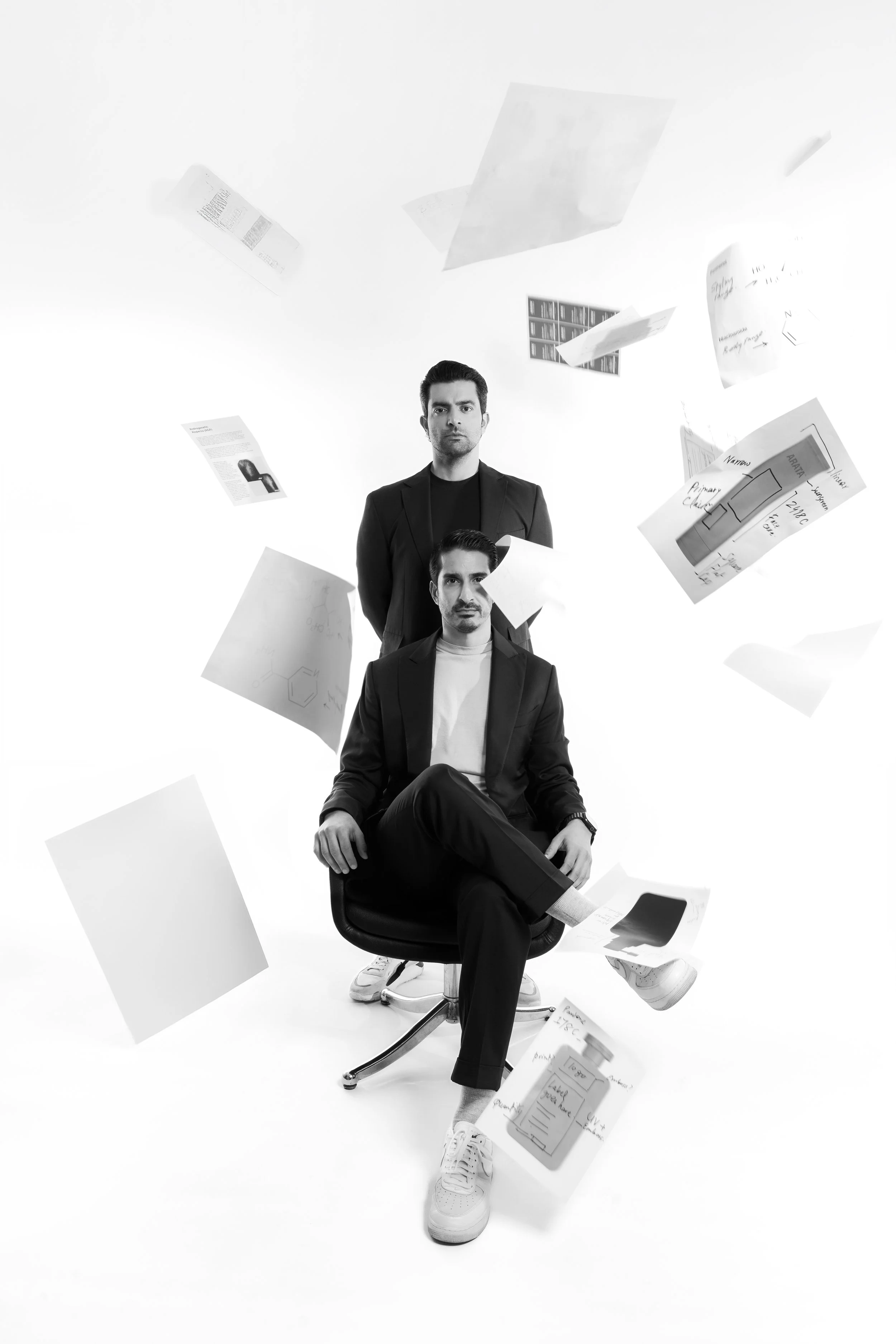 Two men in black suits and white sneakers are in a white studio with papers flying around them. One man is sitting on a chair with crossed legs, while the other stands behind him. The papers contain images and text, some handwritten notes.