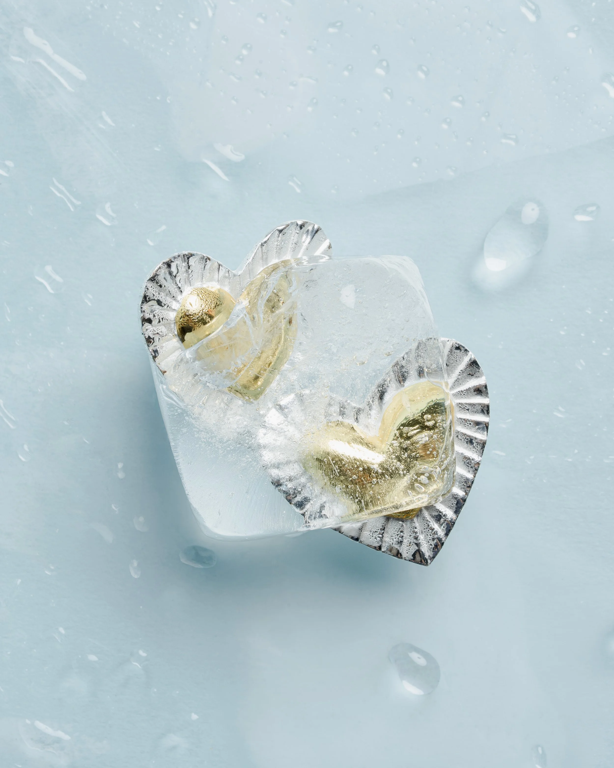 Close-up of a crystal glass with a gold-rimmed slice of lemon inside, placed on a reflective silver coaster, with water droplets on the glass and ice cubes nearby on a light blue surface.