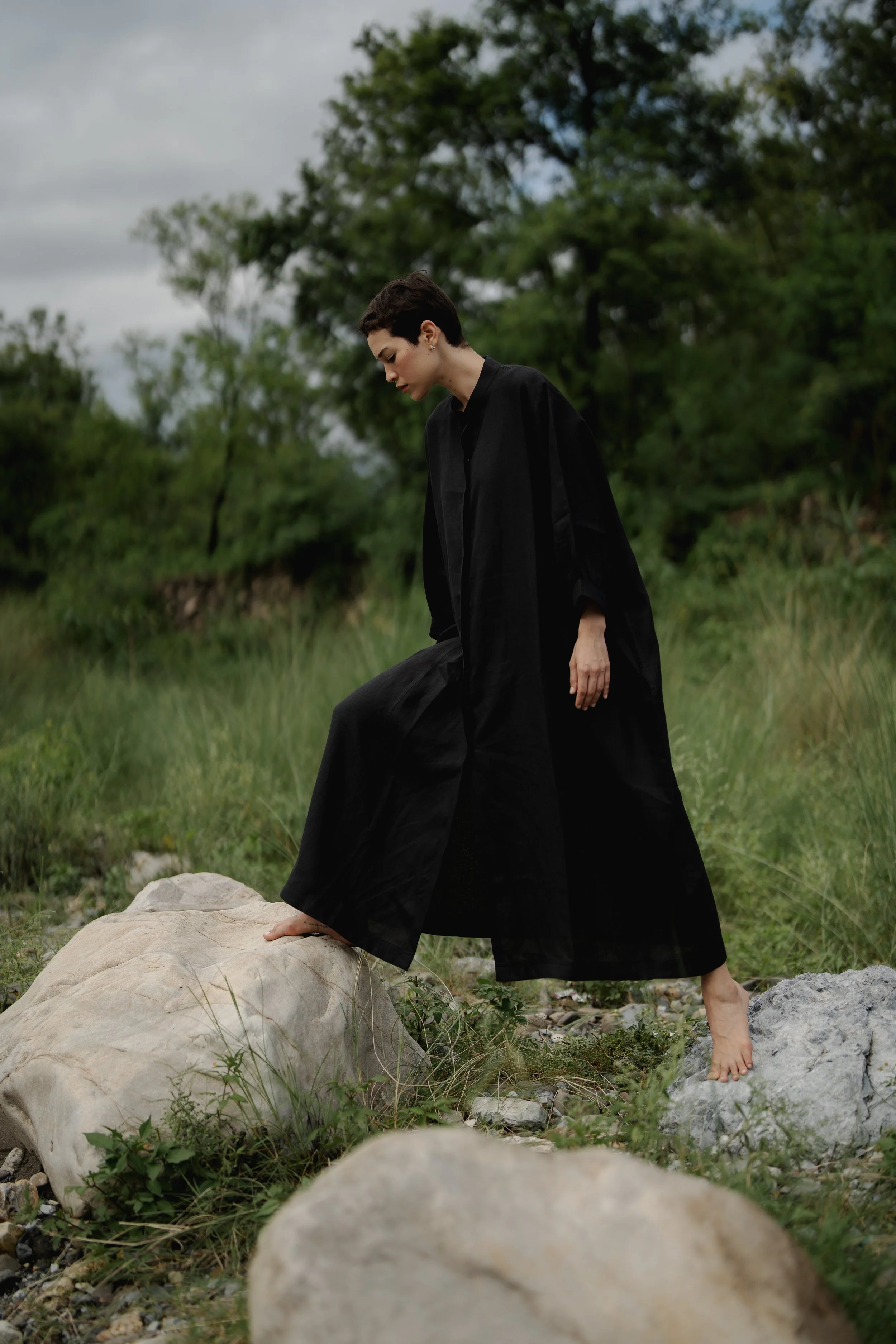 A person with short dark hair wearing a long black dress standing outdoors on rocks with greenery and trees in the background, overcast sky.