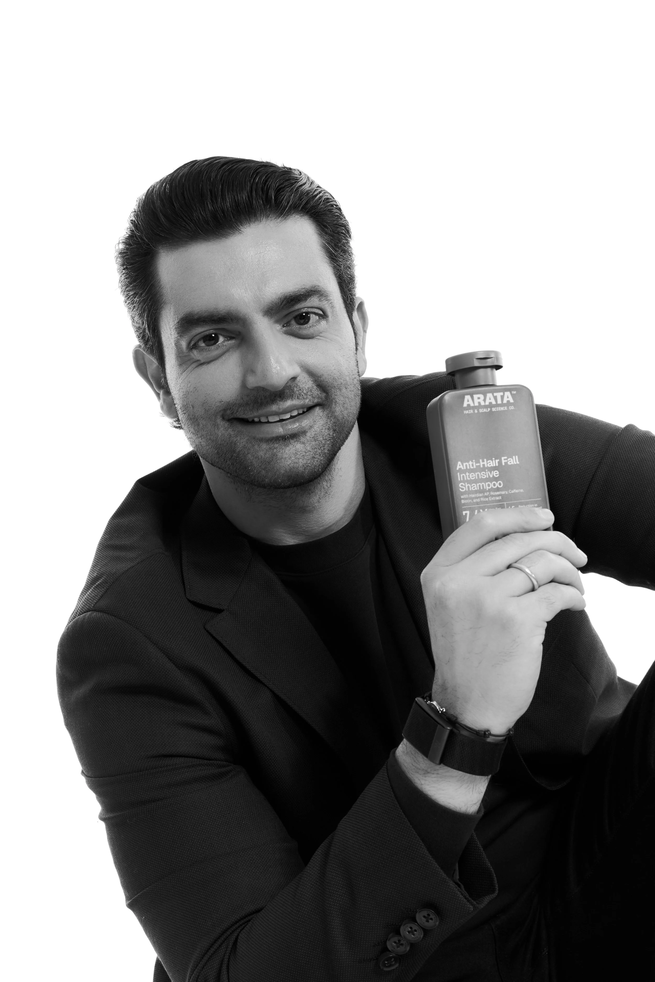 Black and white photo of a smiling man with dark, slicked-back hair, wearing a suit and holding a bottle of Arata anti-hair fall shampoo.