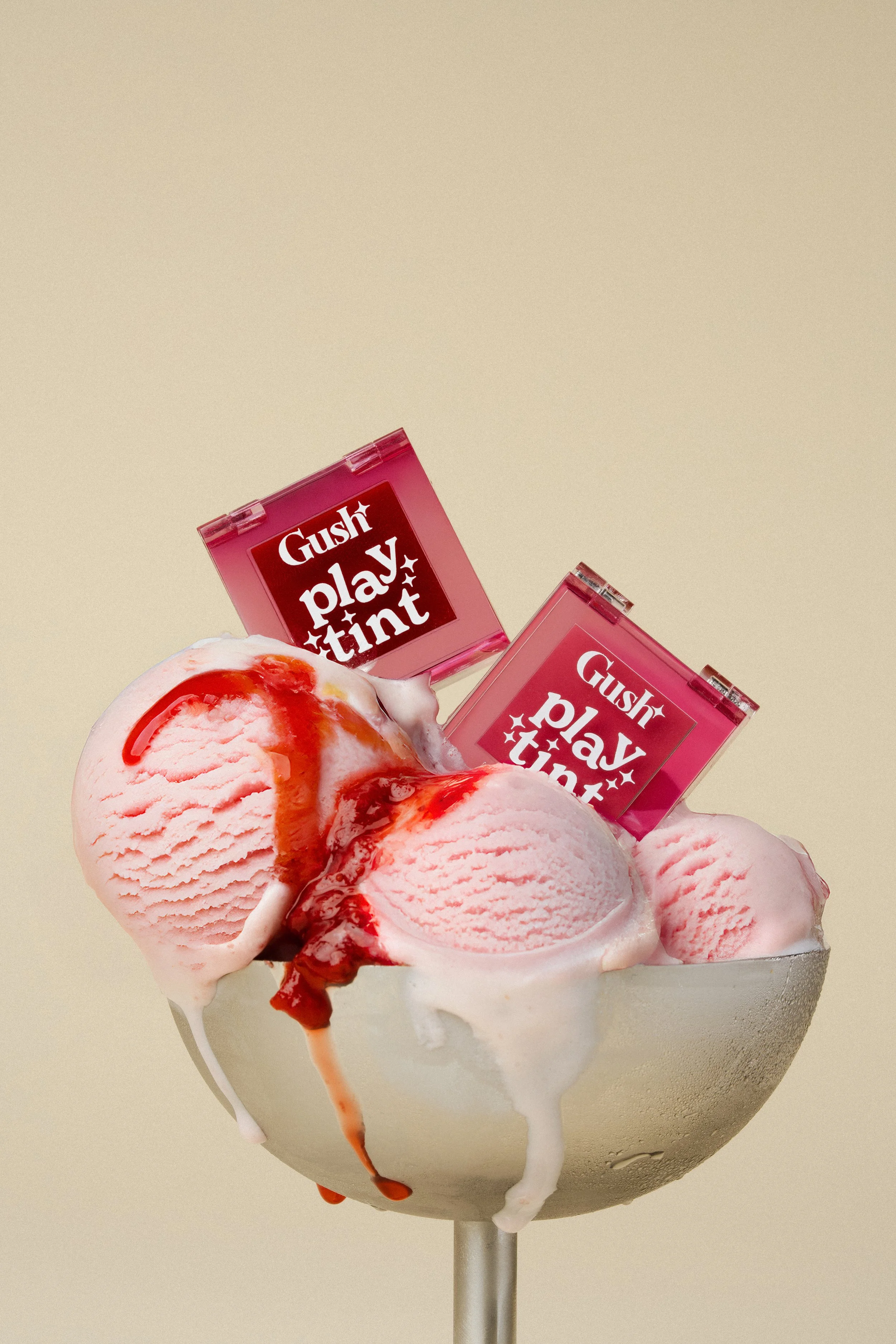 A bowl of strawberry ice cream with red syrup, topped with two pink packaging of Gush Play Tint lip gloss.