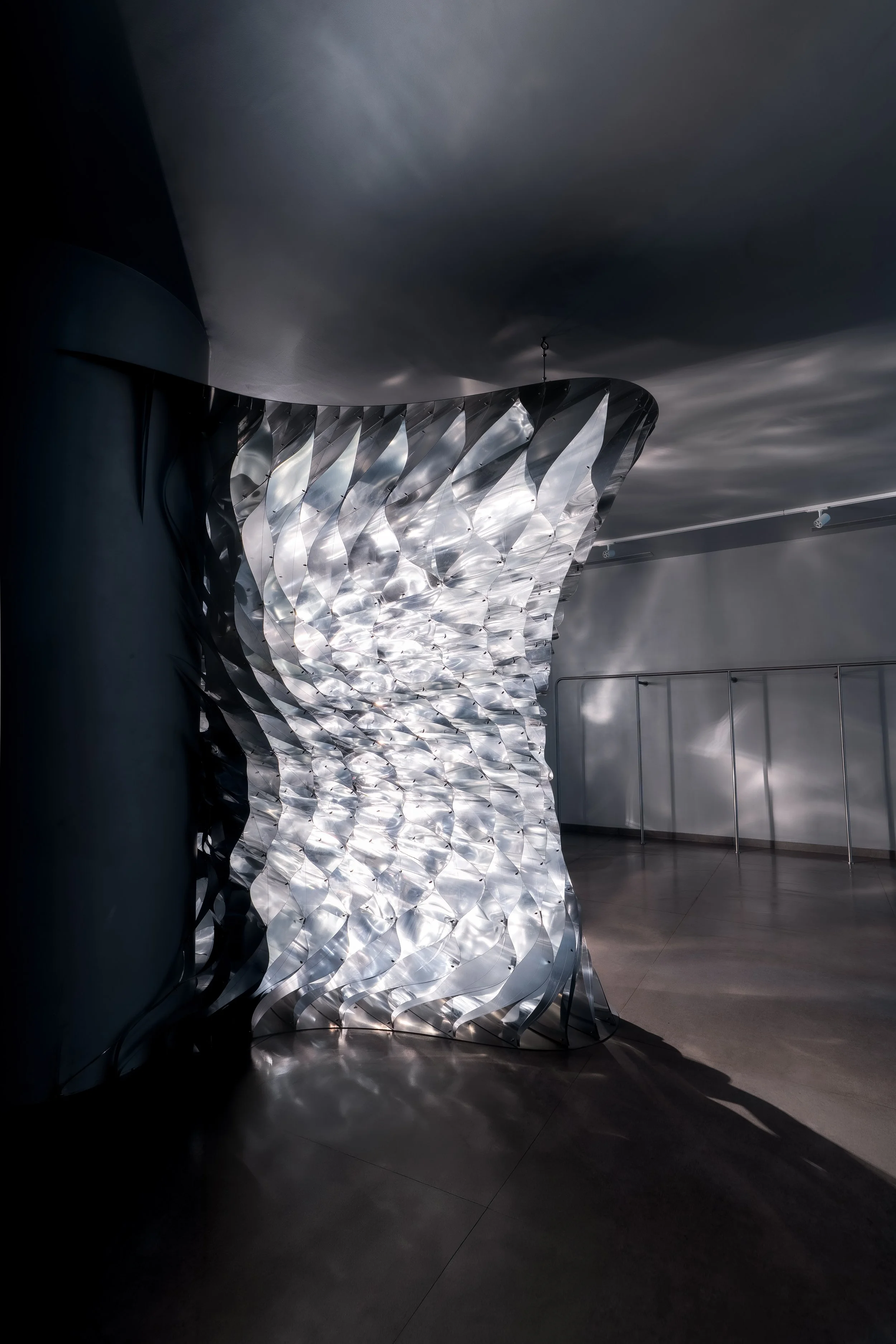 Modern, metallic, wave-like architectural sculpture or installation inside a building with a dark ceiling and gray walls, with sunlight creating shadows on the floor.