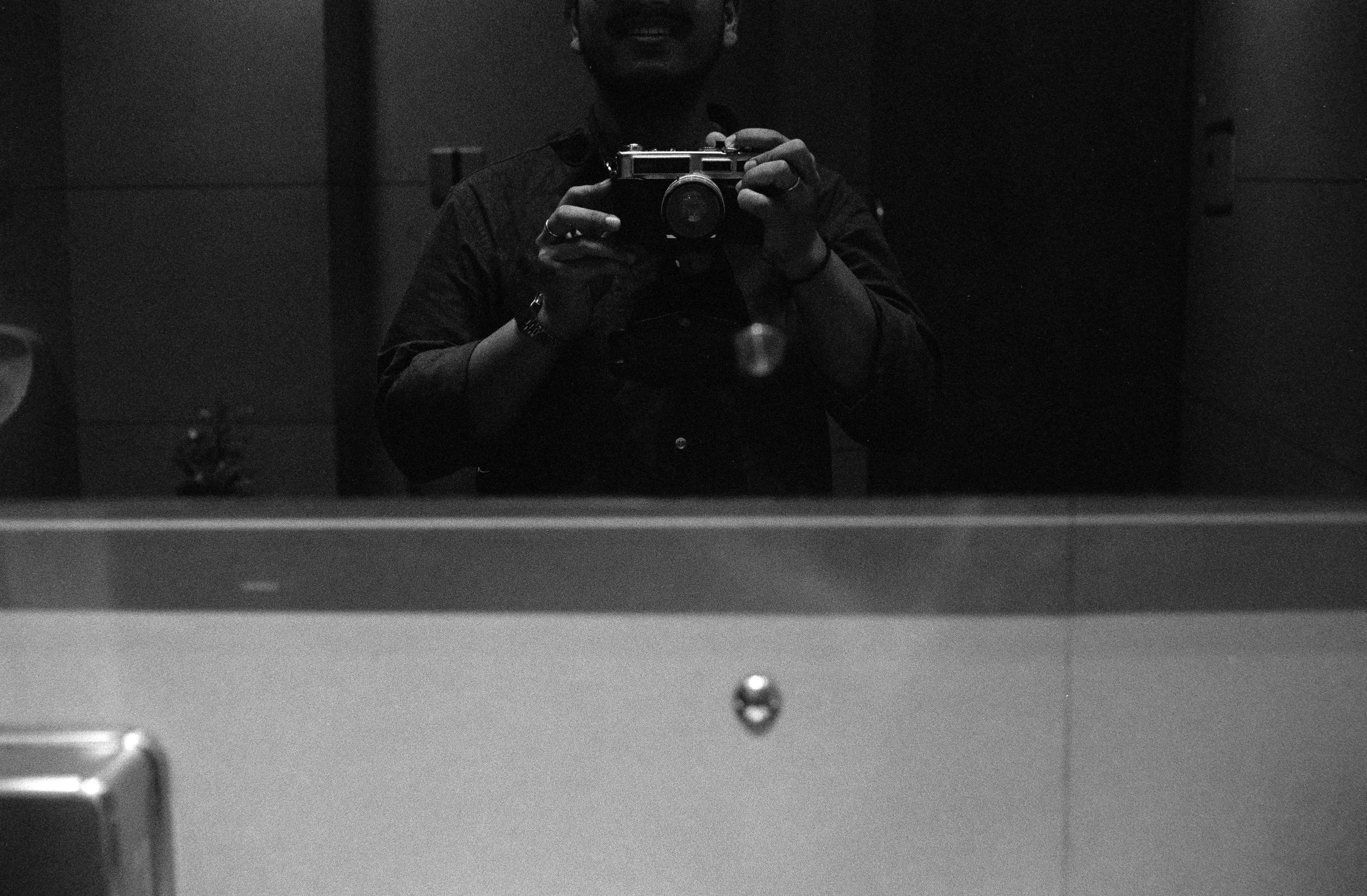 A person taking a selfie in a mirror, holding a camera. The photo is in black and white, with the person wearing glasses and a watch.