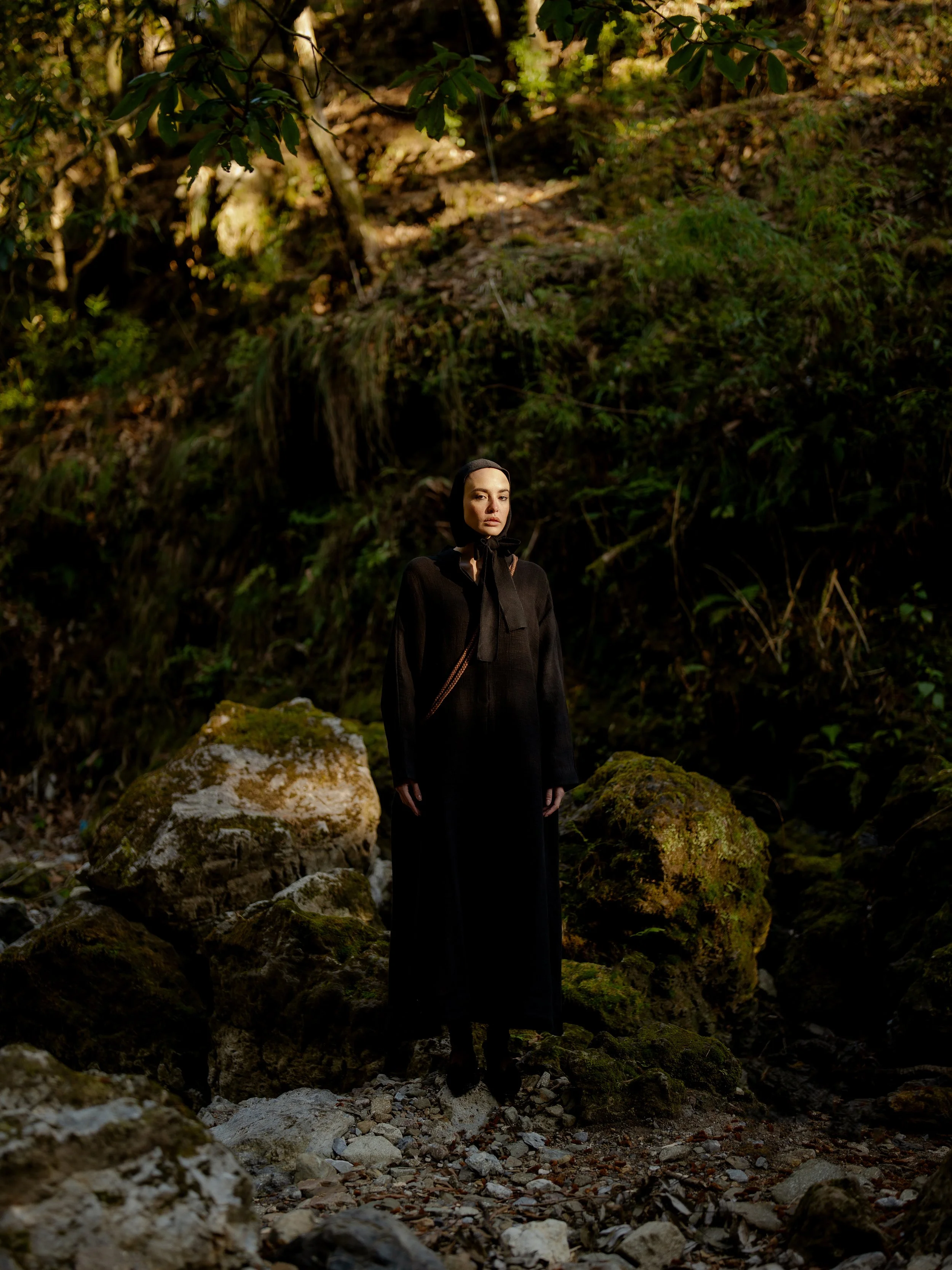 A woman with dark hair and a black outfit standing among rocks in a forested area with trees and moss in the background.
