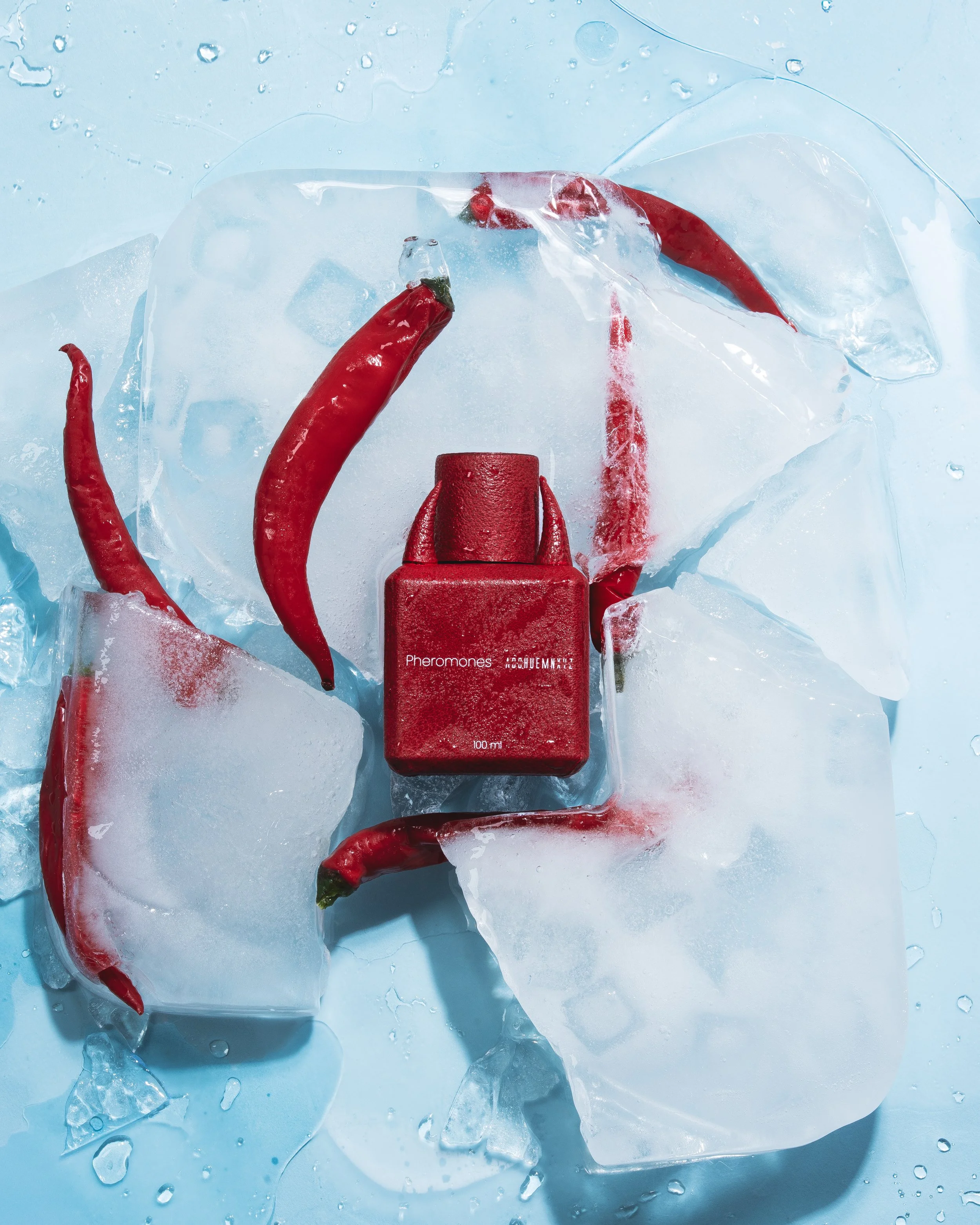 Red chili peppers and a red perfume bottle labeled "Pheromones" on ice cubes with water droplets on a blue background.