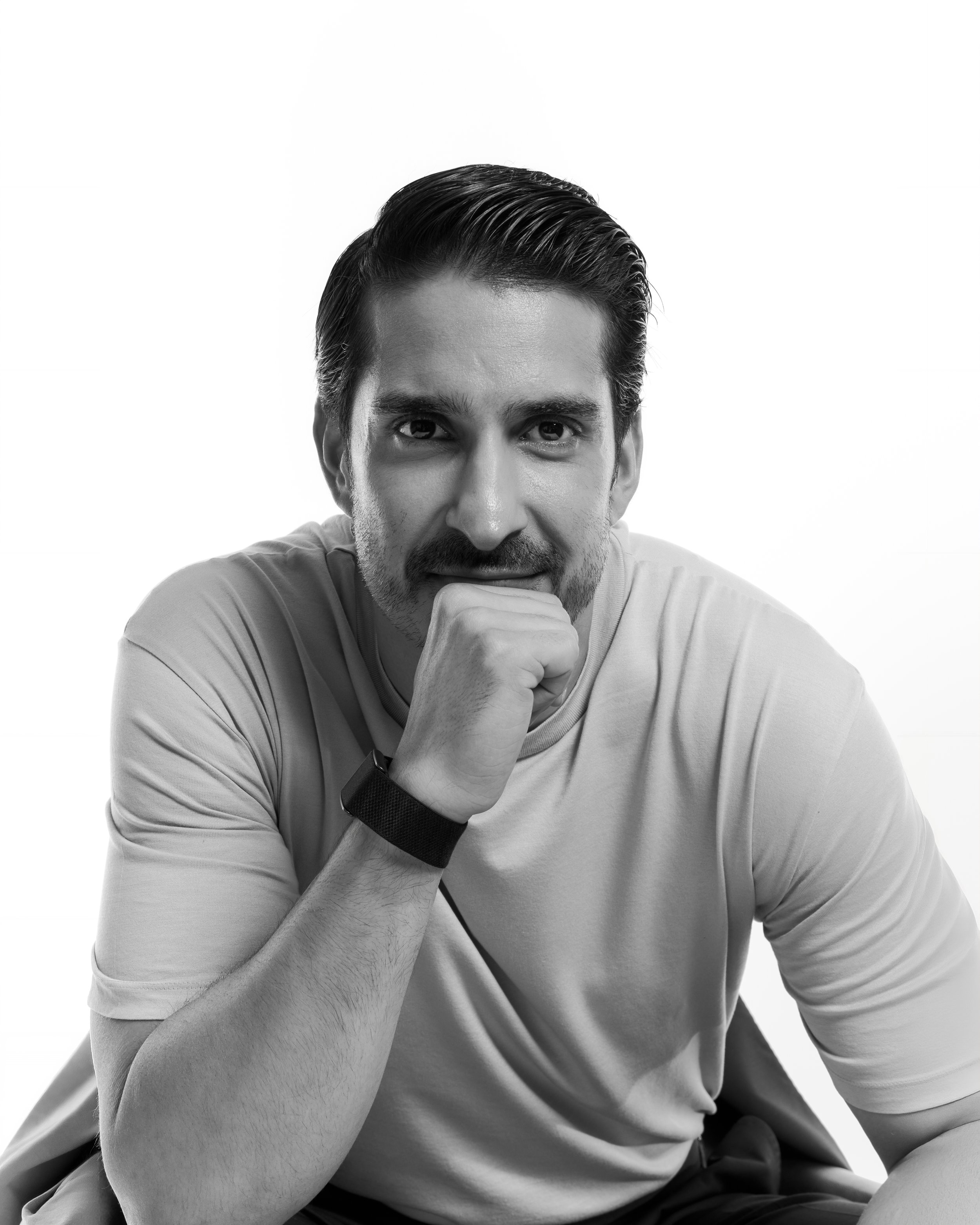 A black and white photo of a man with dark hair styled backwards, wearing a light-colored shirt. He is resting his chin on his right hand, gazing directly at the camera with a contemplative expression.