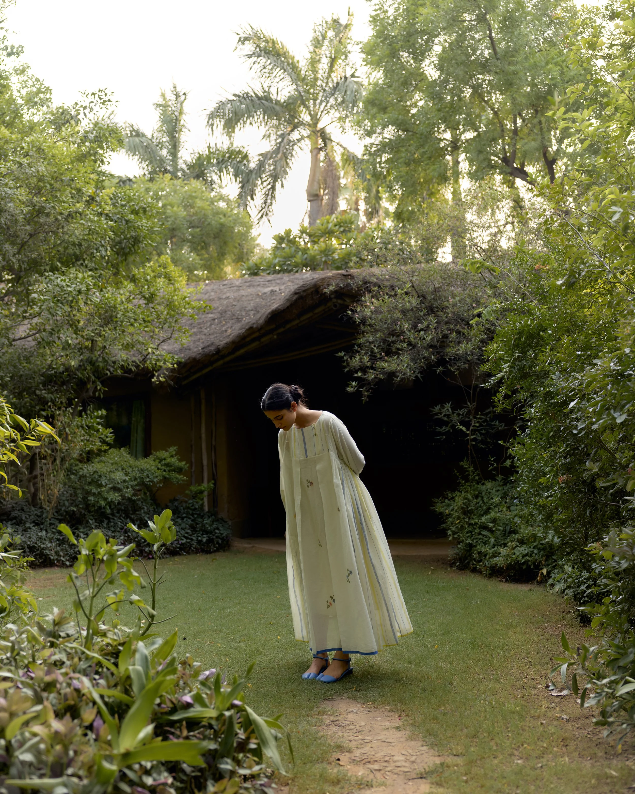A woman wearing a long yellow dress with floral details, blue shoes, and sunglasses standing on a small dirt path in a lush garden with green plants and trees, and a thatched-roof structure in the background during daytime.