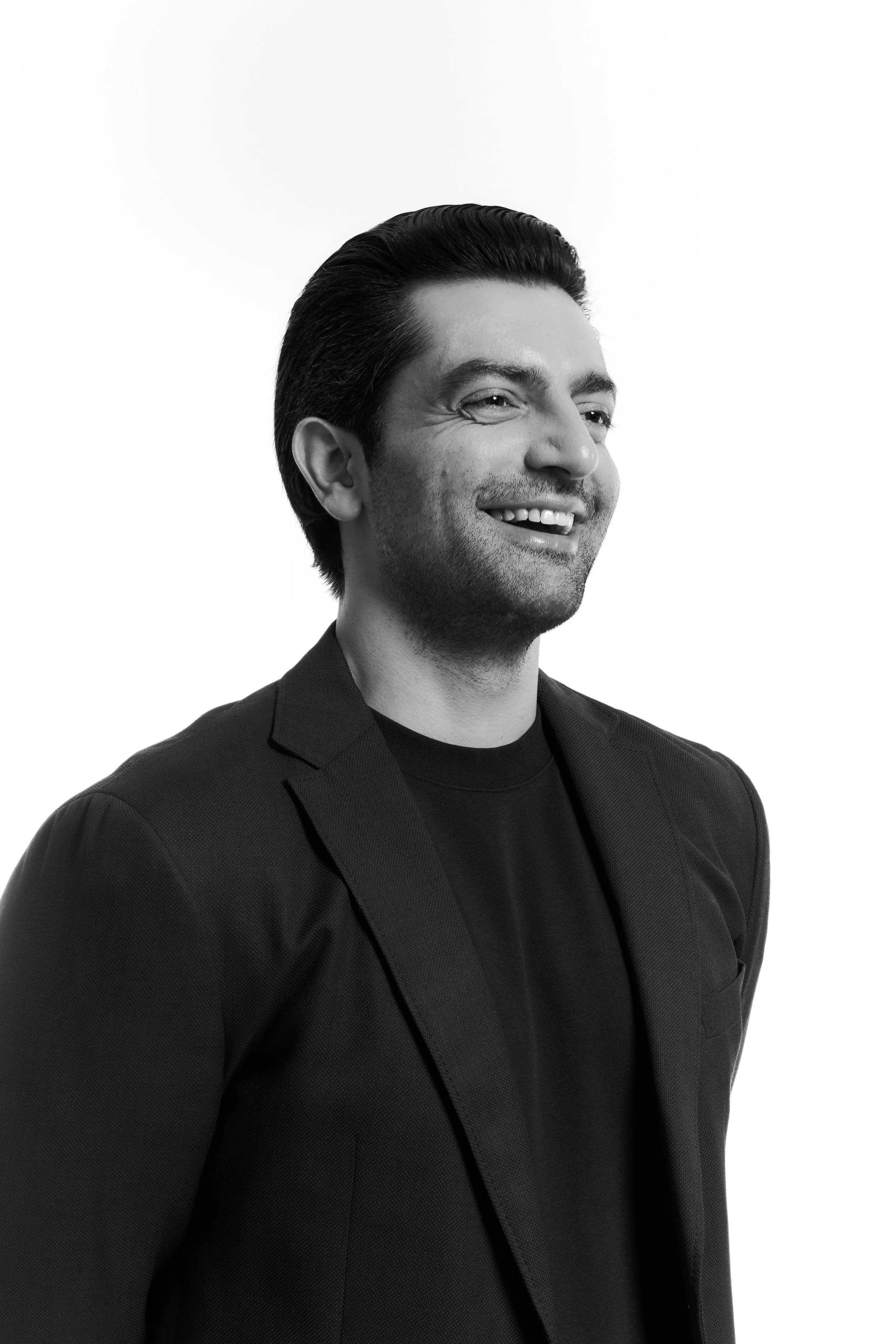 Black and white portrait of a man smiling, wearing a dark blazer and t-shirt, with slicked-back hair, on a plain white background.
