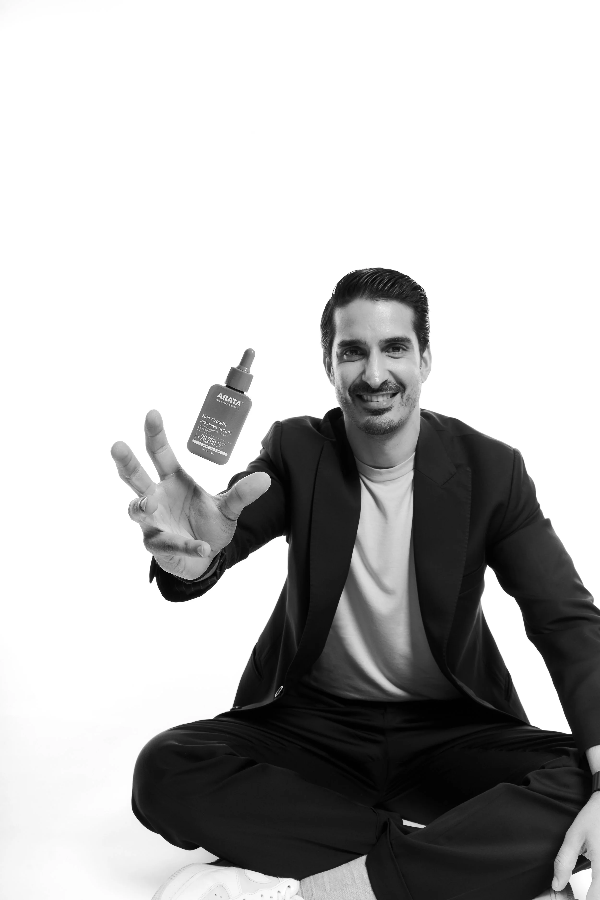 Black and white photo of a smiling man sitting in a cross-legged position, wearing a suit jacket, t-shirt, and sneakers, holding a bottle of Arata hair growth serum.