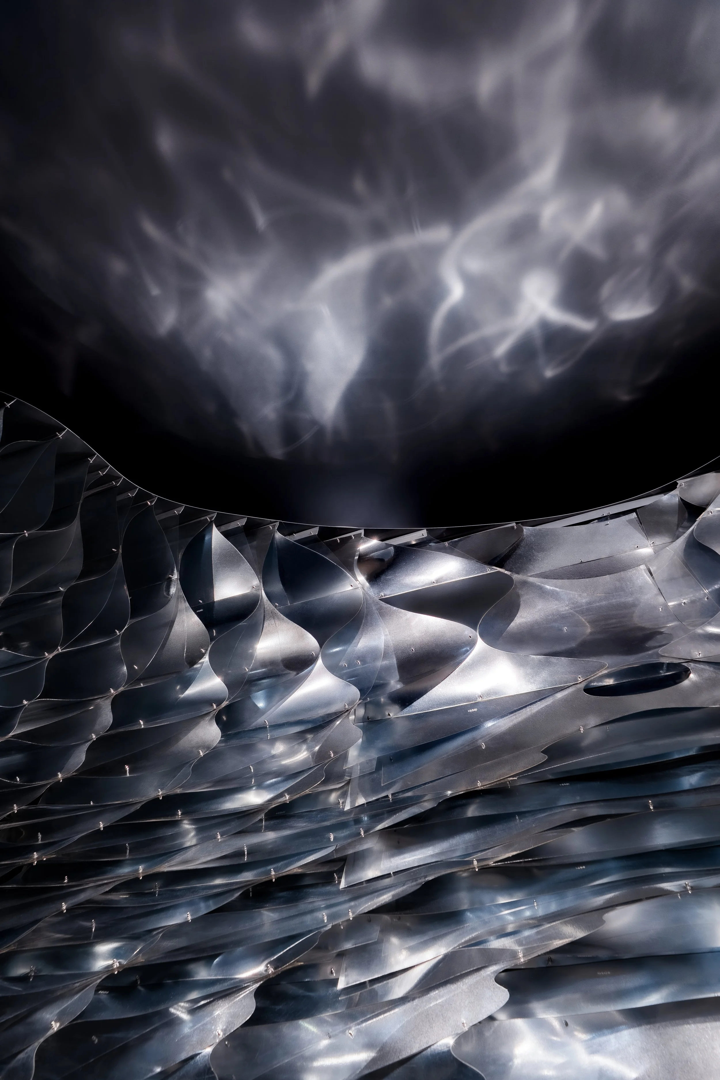 Close-up of a metallic, textured surface with curved, layered elements and a reflection of light creating a shimmering effect.