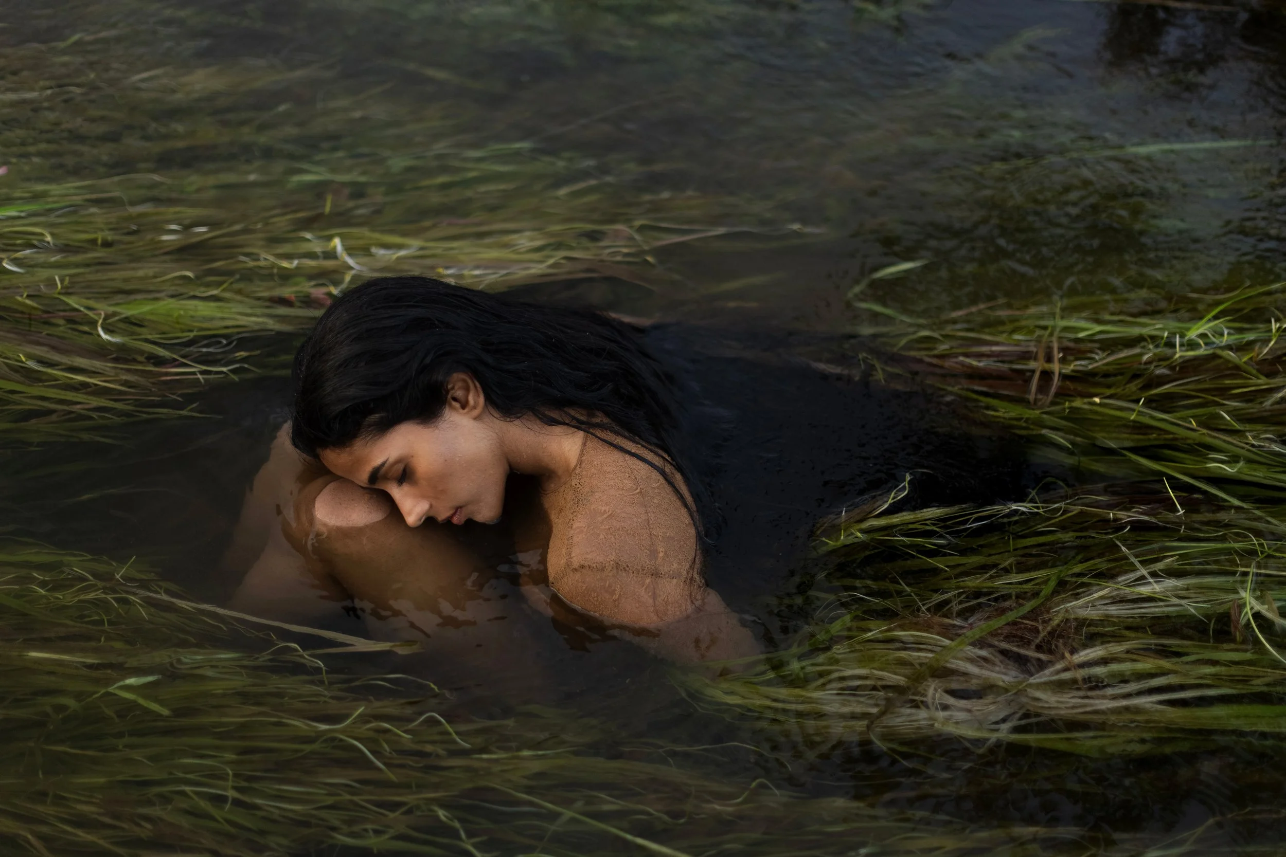 A woman with long dark hair lying in water with green aquatic plants, her eyes closed, and her face resting on her arms.