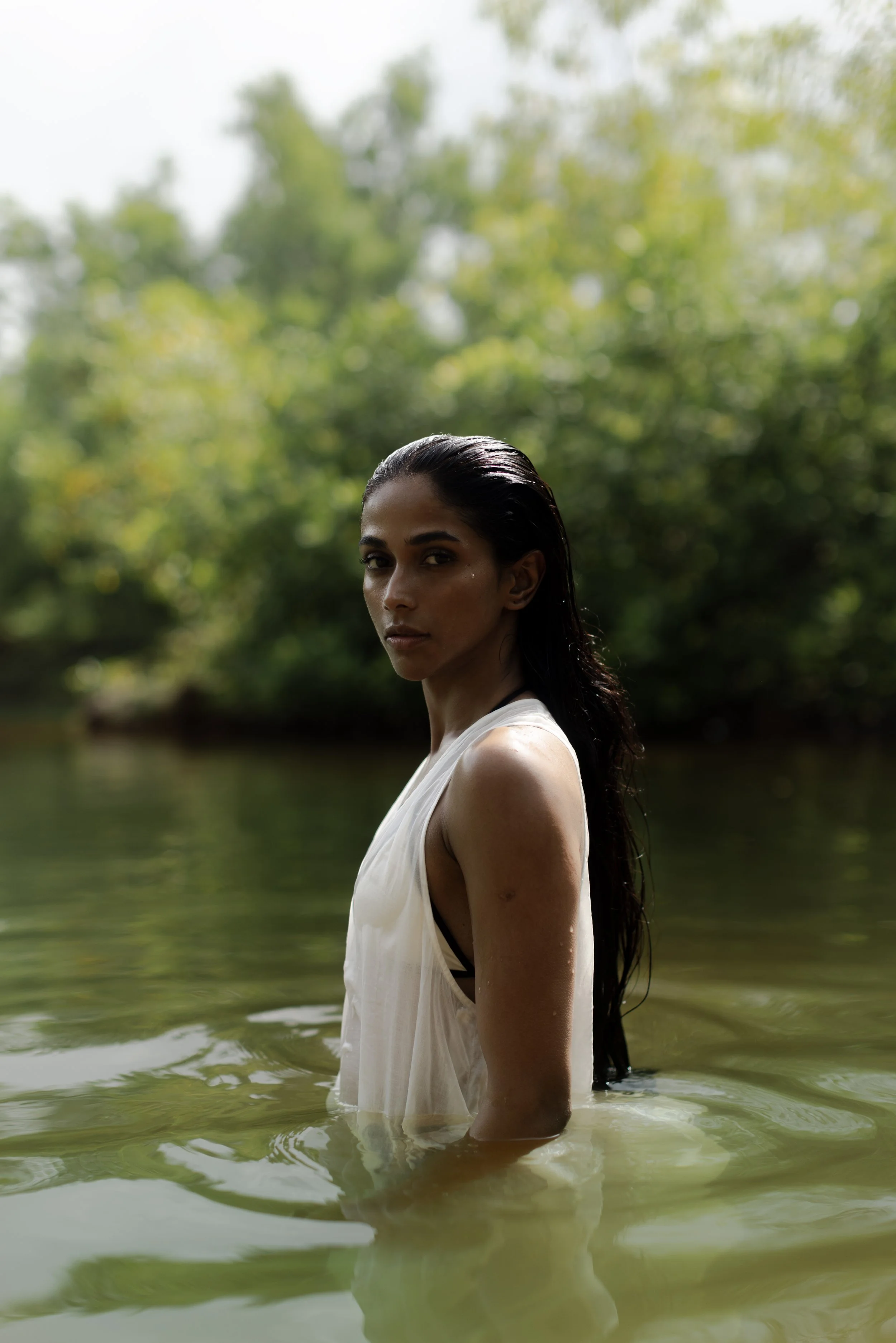 A woman with wet, dark hair stands waist-deep in a body of water, with trees and greenery in the background.