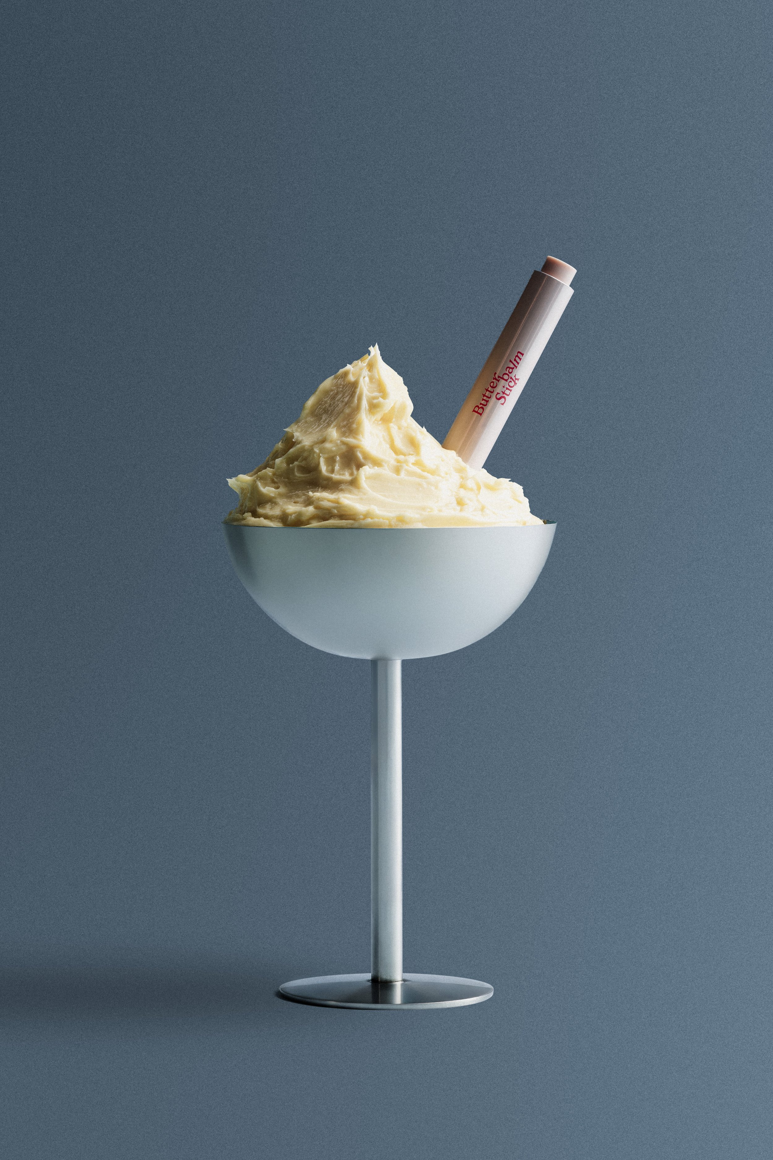 A white bowl filled with a large mound of yellow mashed potatoes, with a tube of Butter Balm Stick inserted into the mashed potatoes, against a blue background.