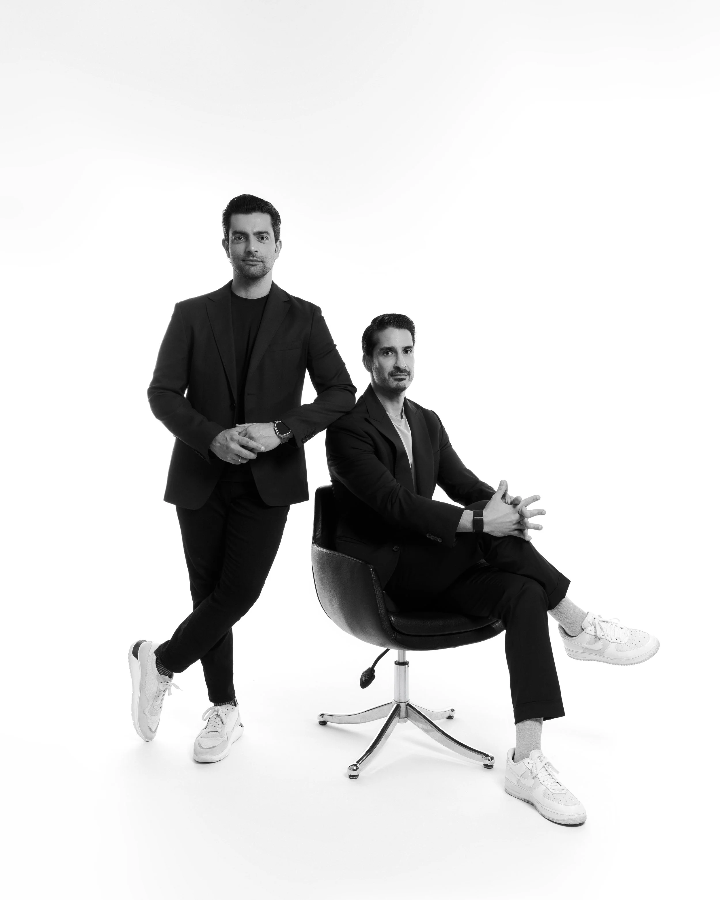 Two men with dark hair in black suits and white sneakers. One man is standing confidently with arms crossed, and the other is sitting on a swivel chair with one leg crossed over the other. Both have serious expressions. The background is plain white.