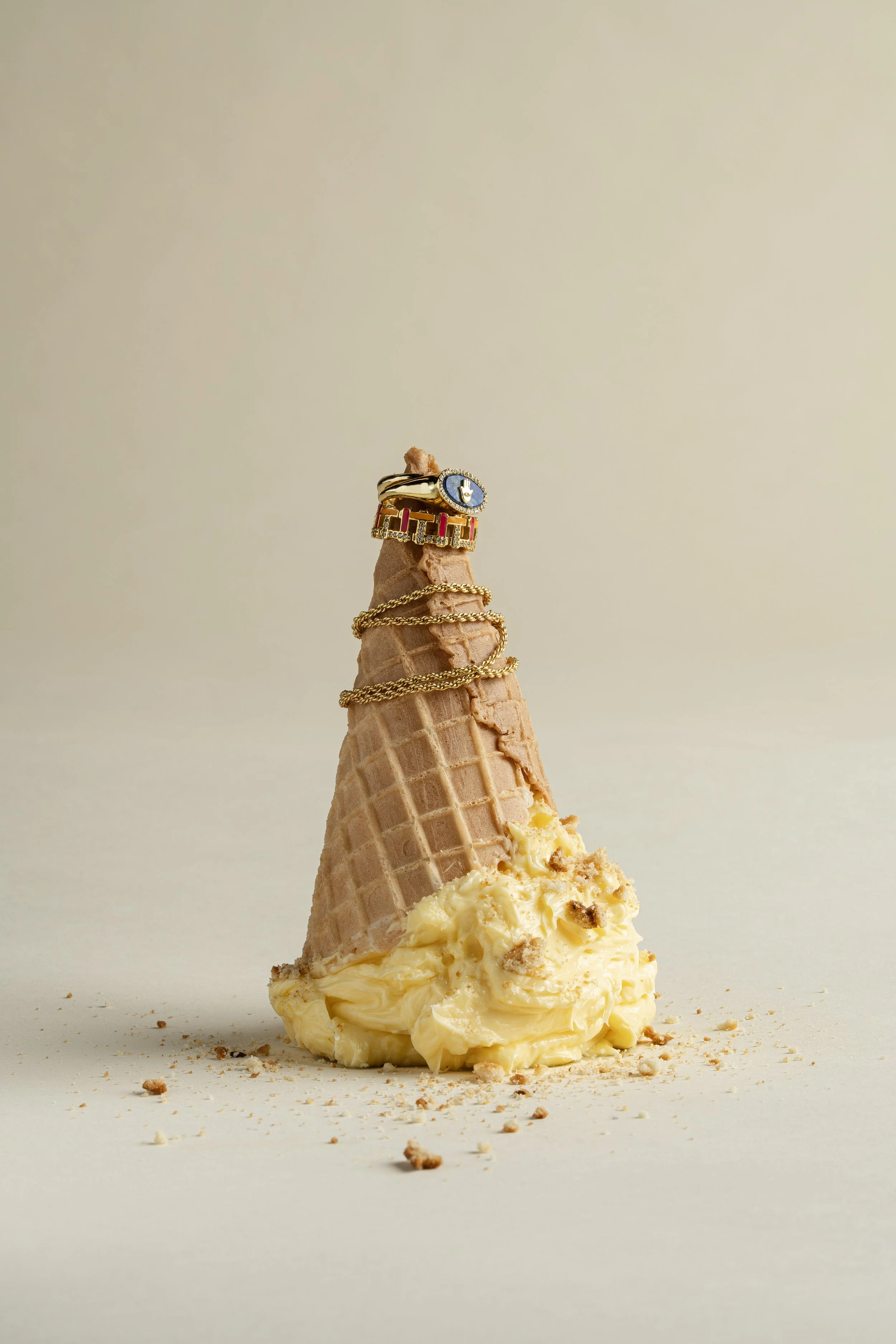 Ice cream cone with caramel, gold rings with jewelry, and a ring with a blue gemstone on top on a plain background