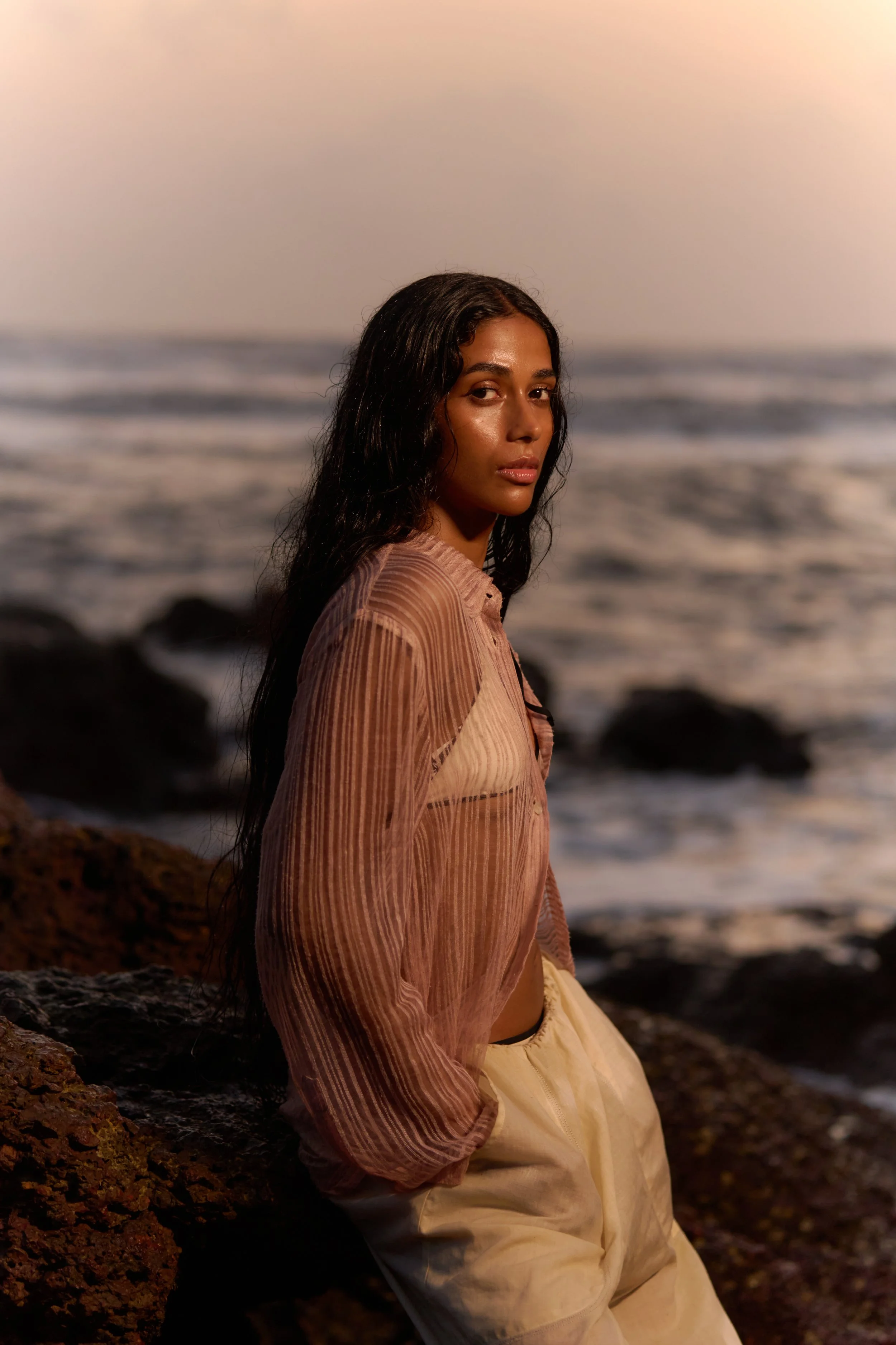 A woman with long dark hair sitting on rocks near the ocean at sunset, wearing a sheer pinkish-brown top and cream-colored pants.