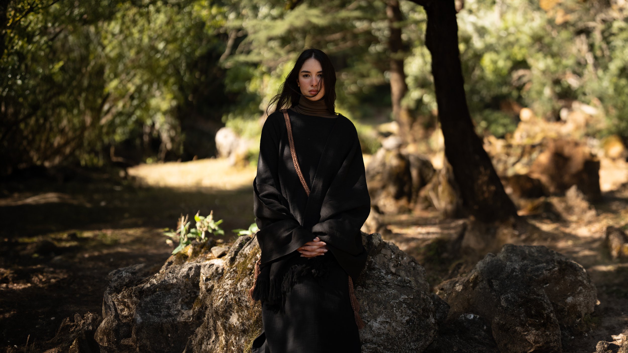 A young woman with dark hair, wearing dark clothing, sitting on a large rock in a forest with trees and sunlight filtering through the leaves.