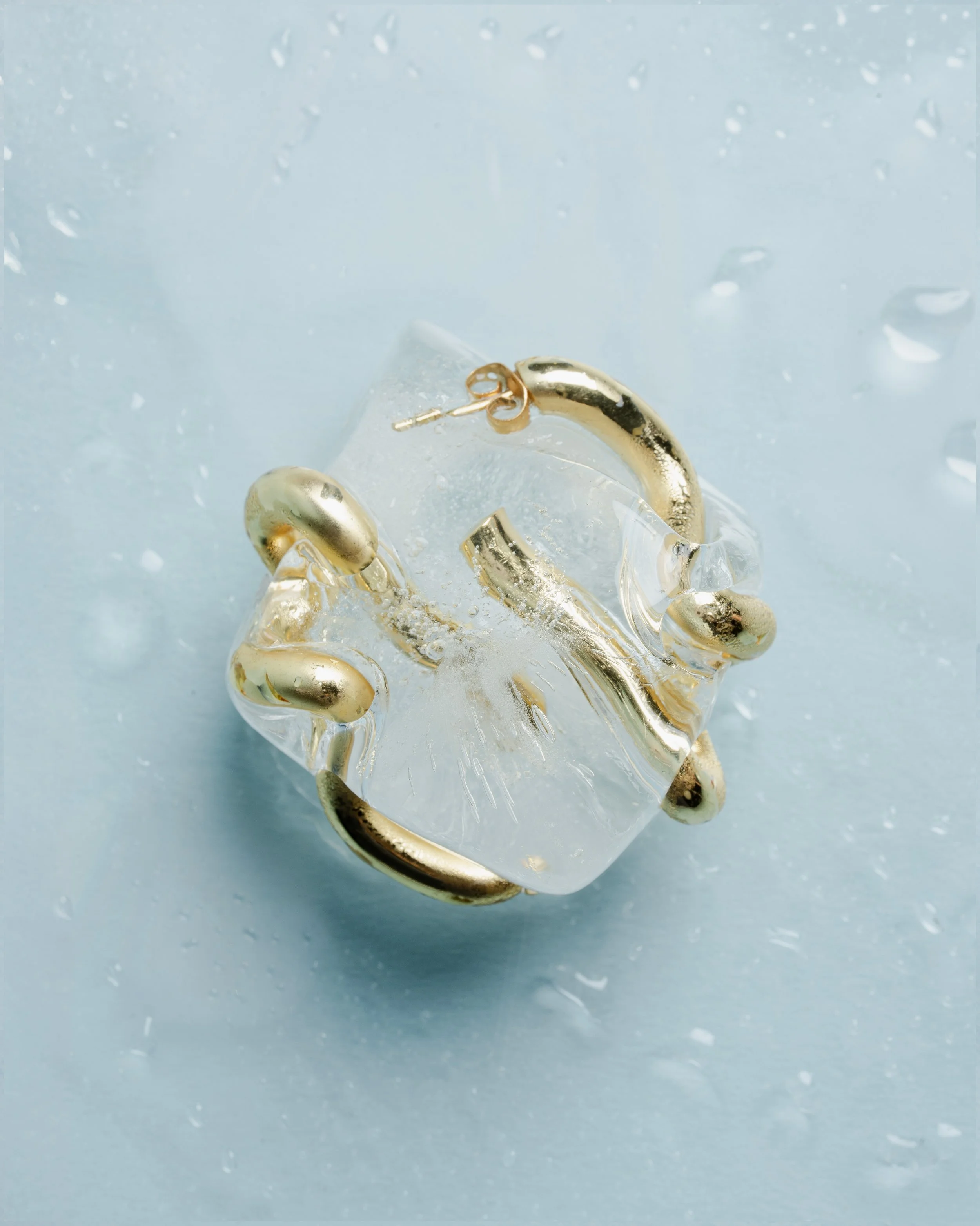 Close-up of a gold and clear jewelry piece, possibly a bracelet or ring, resting on a blue surface with water droplets.