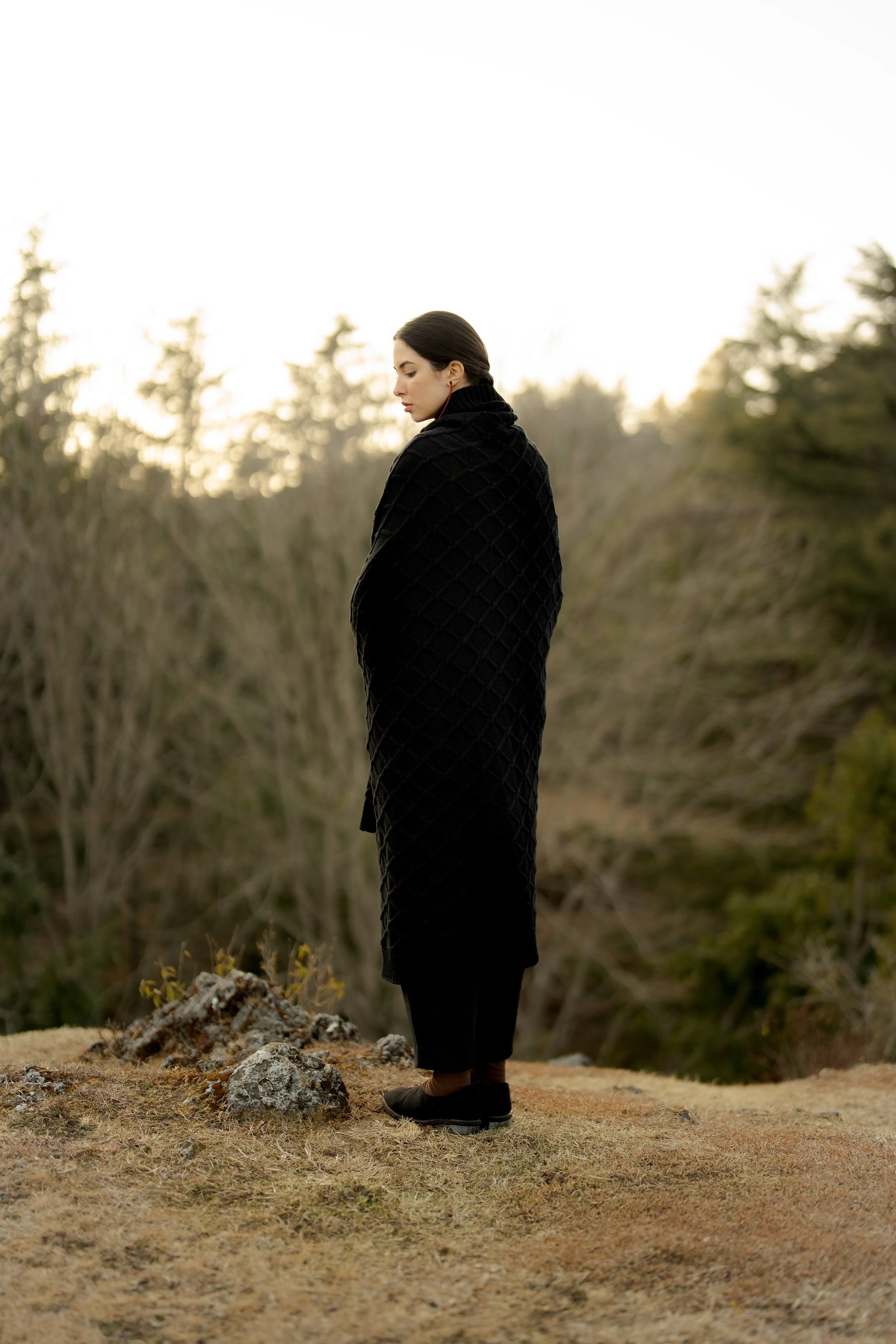 A woman dressed in black with dark hair, standing outdoors on rocky ground, surrounded by sparse grass and bushes, with trees in the background and a bright sky.