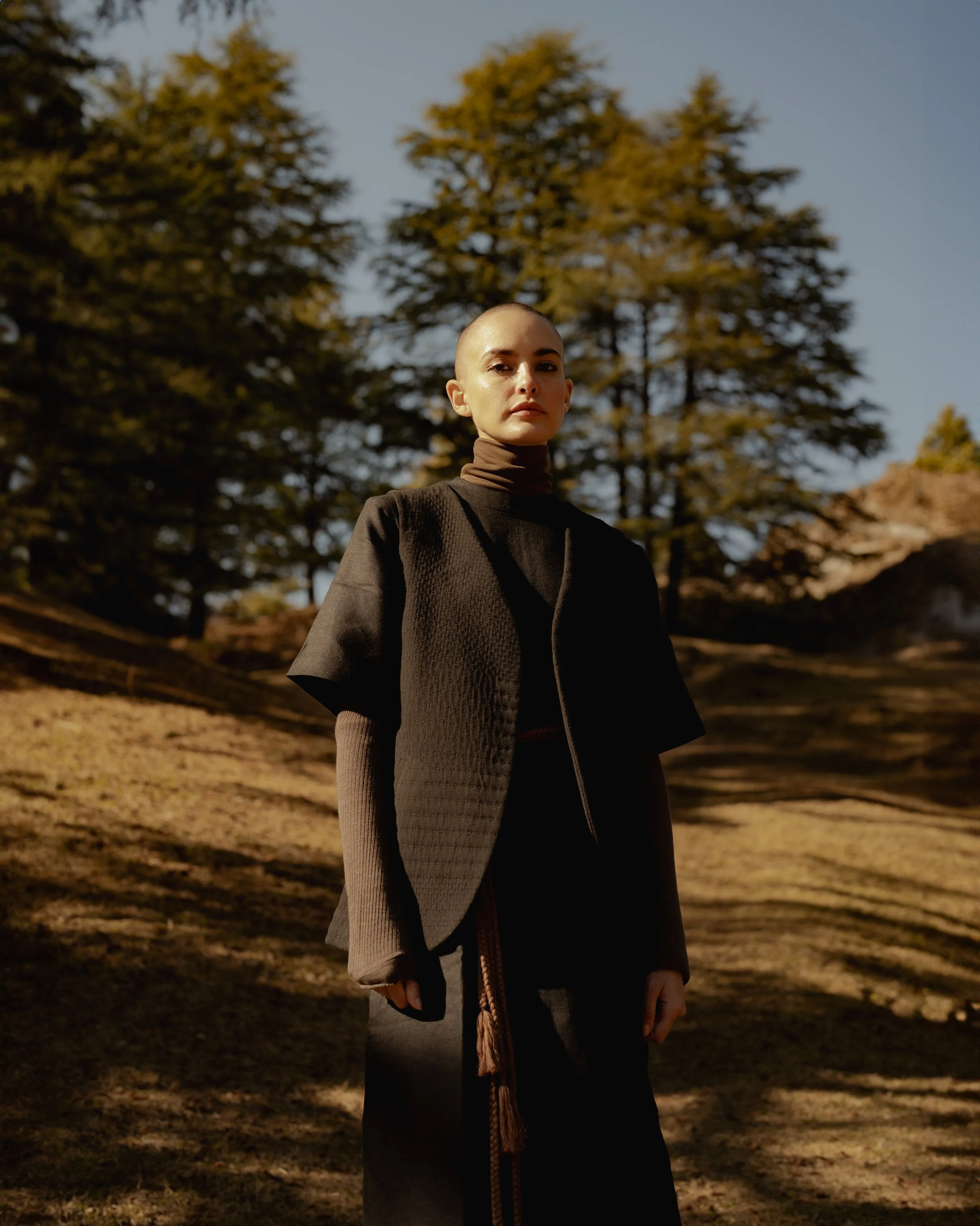 A woman with a shaved head standing outdoors in a forest with tall trees during late afternoon sunlight.