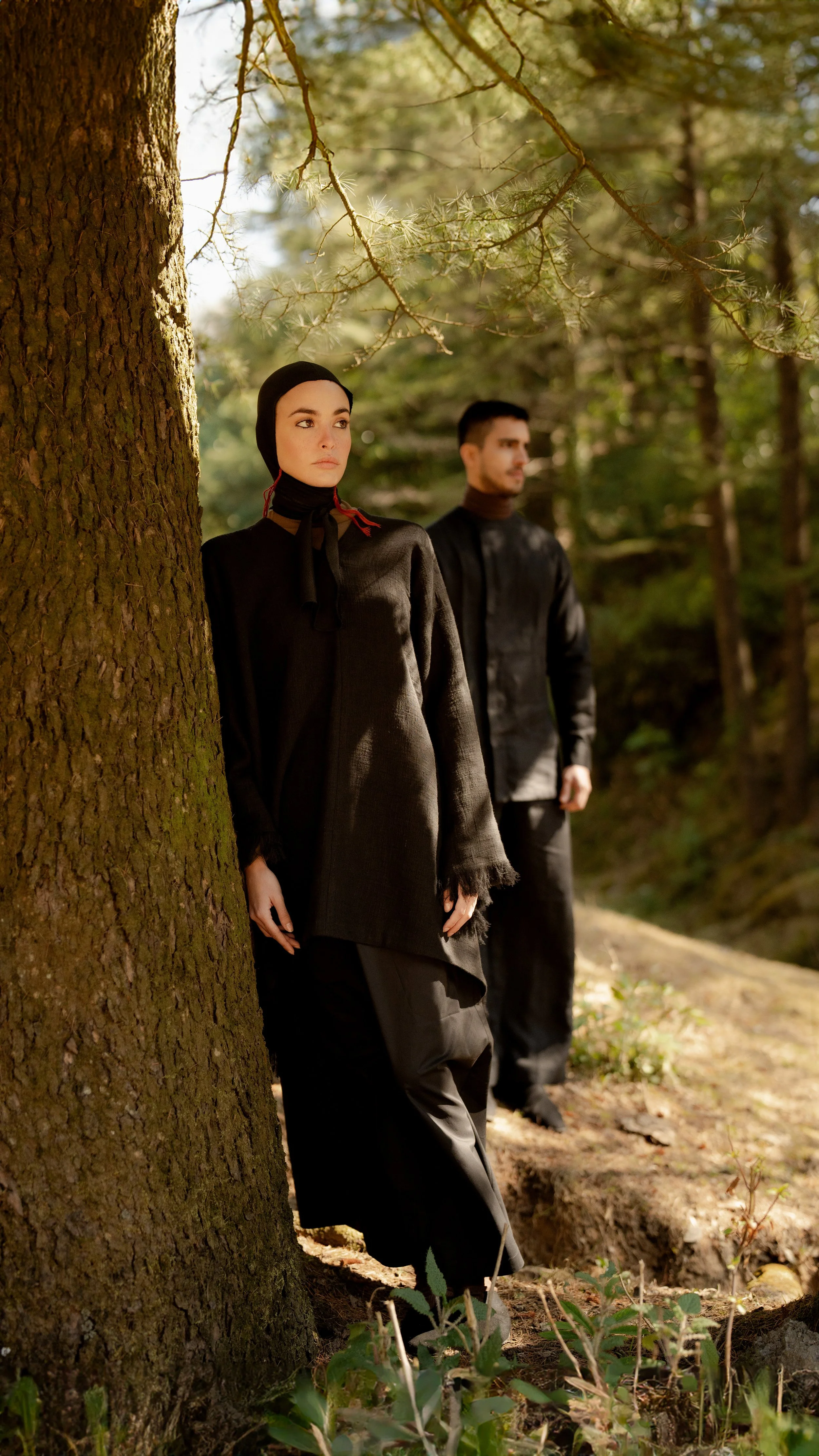 A woman in a black headscarf and long black coat standing behind a tree in a forest, with a man in similar black clothing in the background, blurry and looking away.