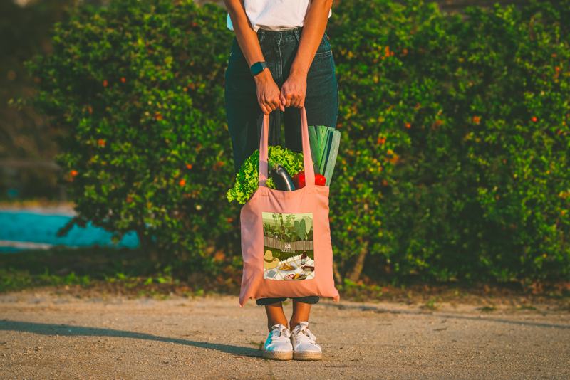 Personne ayant fait un marché forain de la Réunion. Elle porte en extérieur un tote bag rose corail avec une illustration imprimée d'un pique-nique Péi dans un jardin. Le tote bag est rempli de légumes frais et locaux sur fond de végétation.