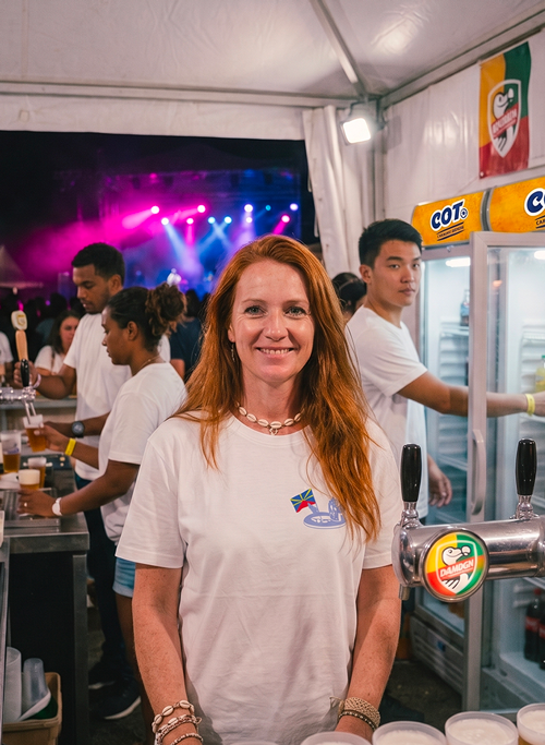 Une bénévole souriante derrière un bar  lors d'un festival ou événement culturel, avec une scène, des lumières colorées, des tireuses à bière et des frigos en arrière-plan. Elle porte un t-shirt personnalisé Tetal'eau studio.