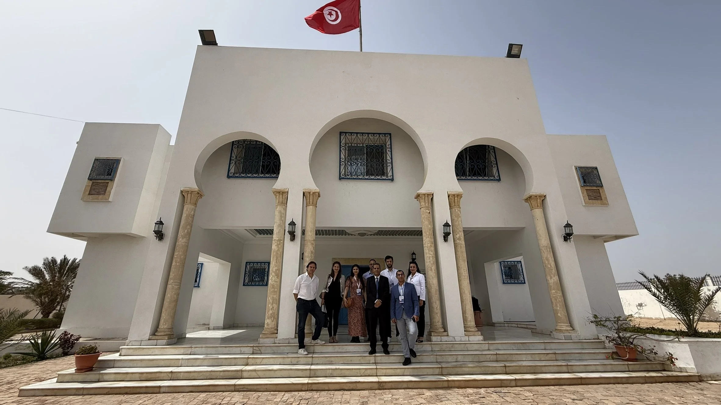Groupe de personnes debout devant un bâtiment blanc avec des colonnes, une façade en forme d'arc et un drapeau tunisien flottant en haut.
