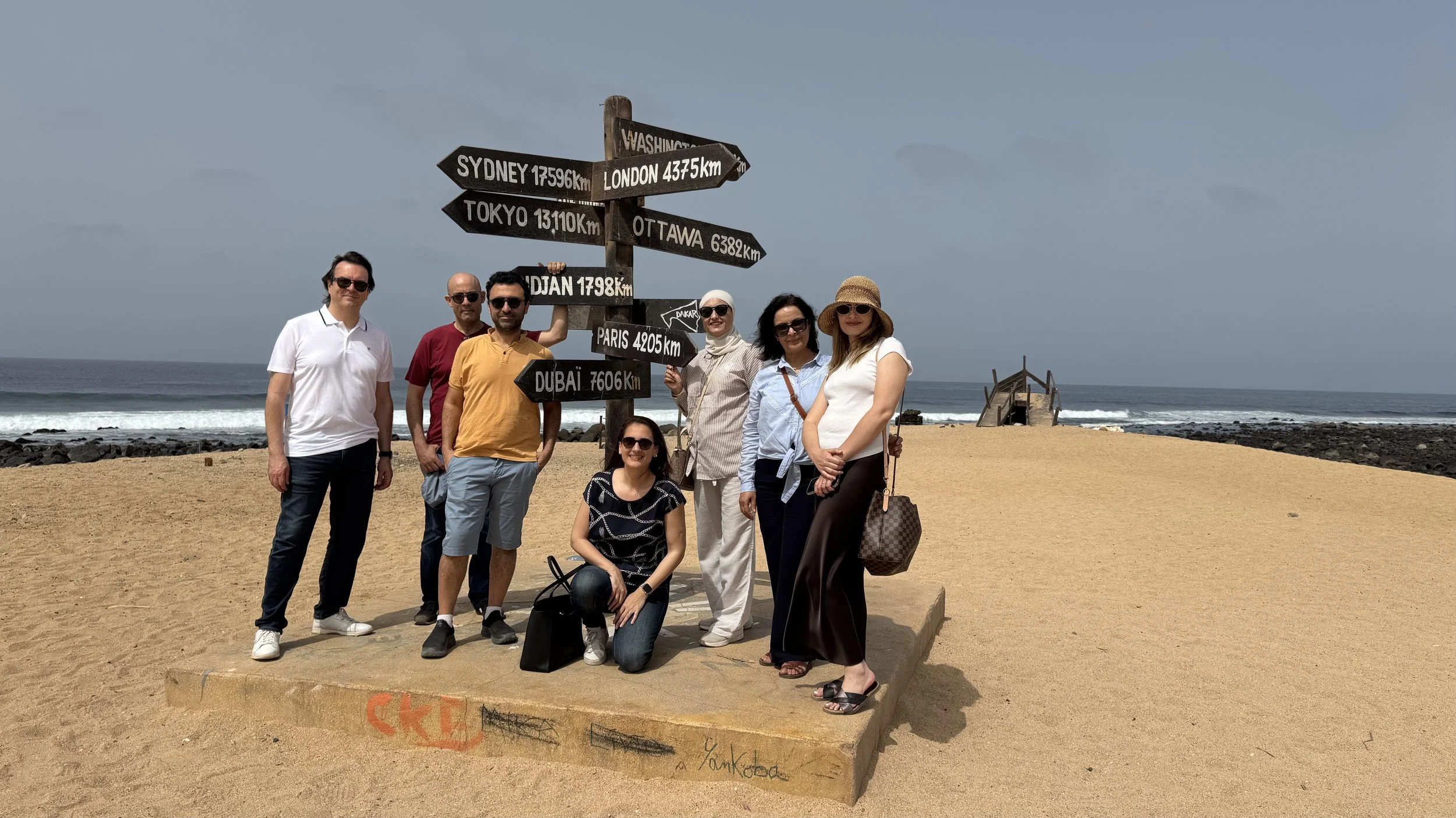 groupe de personnes posant devant un panneau indiquant différentes distances à des villes du monde sur une plage