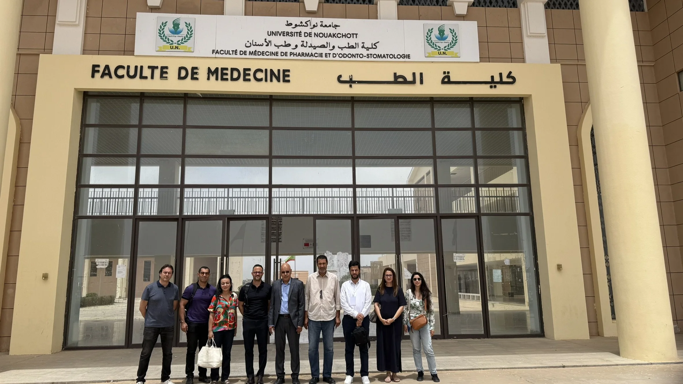 Groupe de personnes debout devant l'entrée de la faculté de médecine à l'Université de Nouakchott, avec un panneau indiquant le nom de l'établissement en arabe, français et arabe, sous un ciel clair.