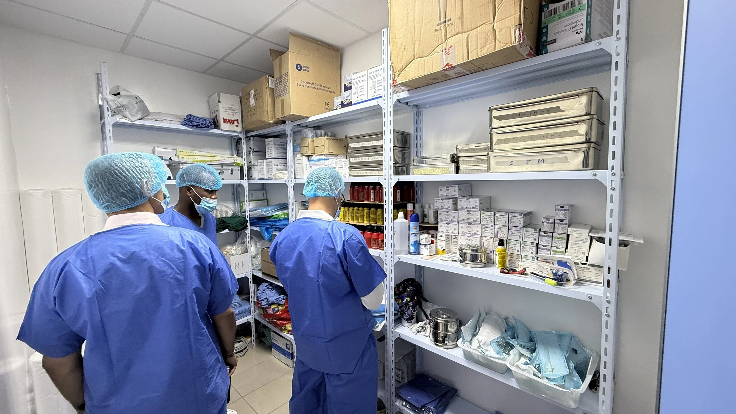 Trois professionnels de la santé portant des uniformes bleus, des charlottes et des masques dans une pièce de stockage de matériel médical, avec des étagères remplies de fournitures et équipements médicaux.
