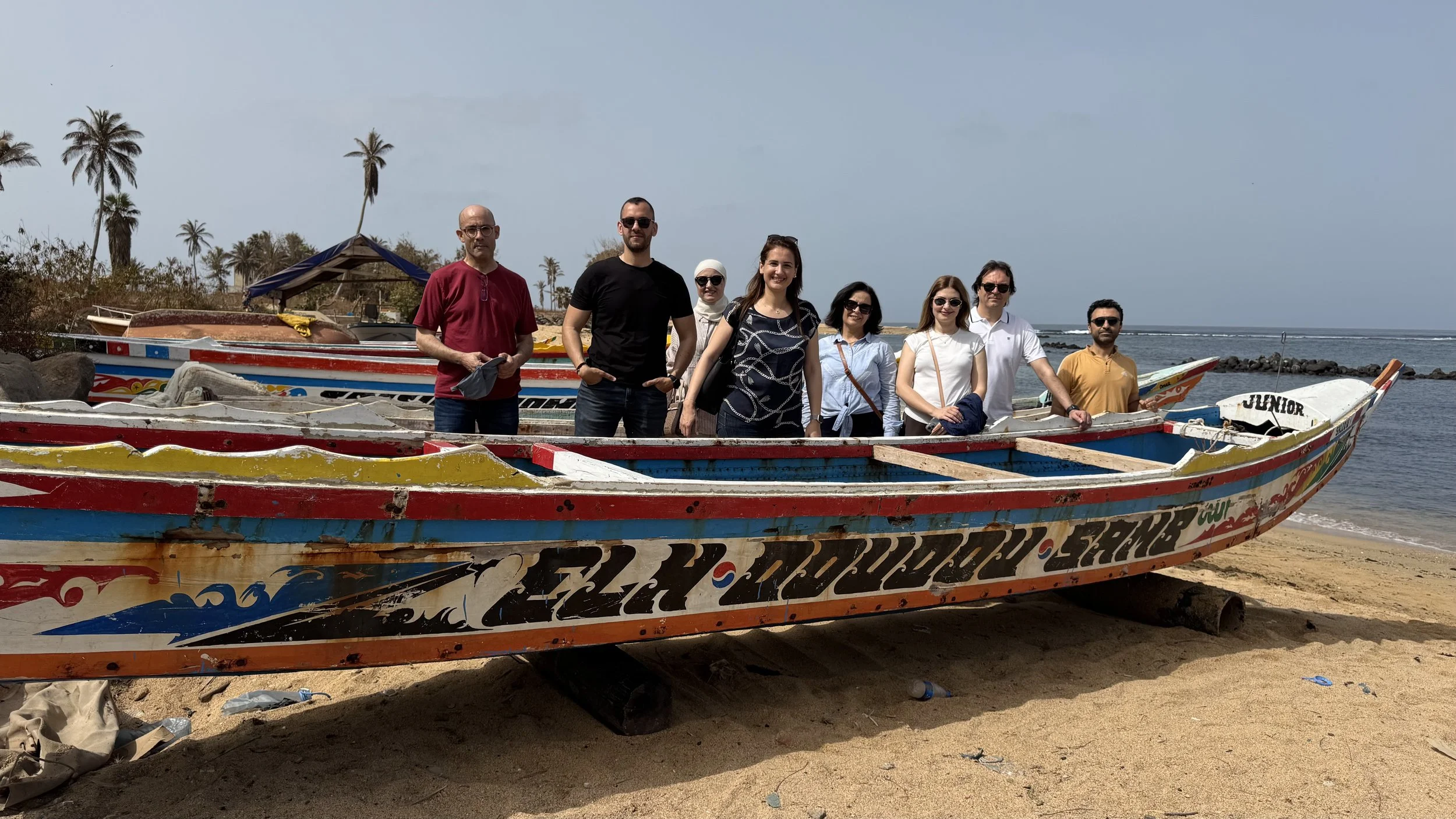 Groupe de huit personnes posant sur une plage avec des bateaux colorés en bois, palmier en arrière-plan et ciel clair.