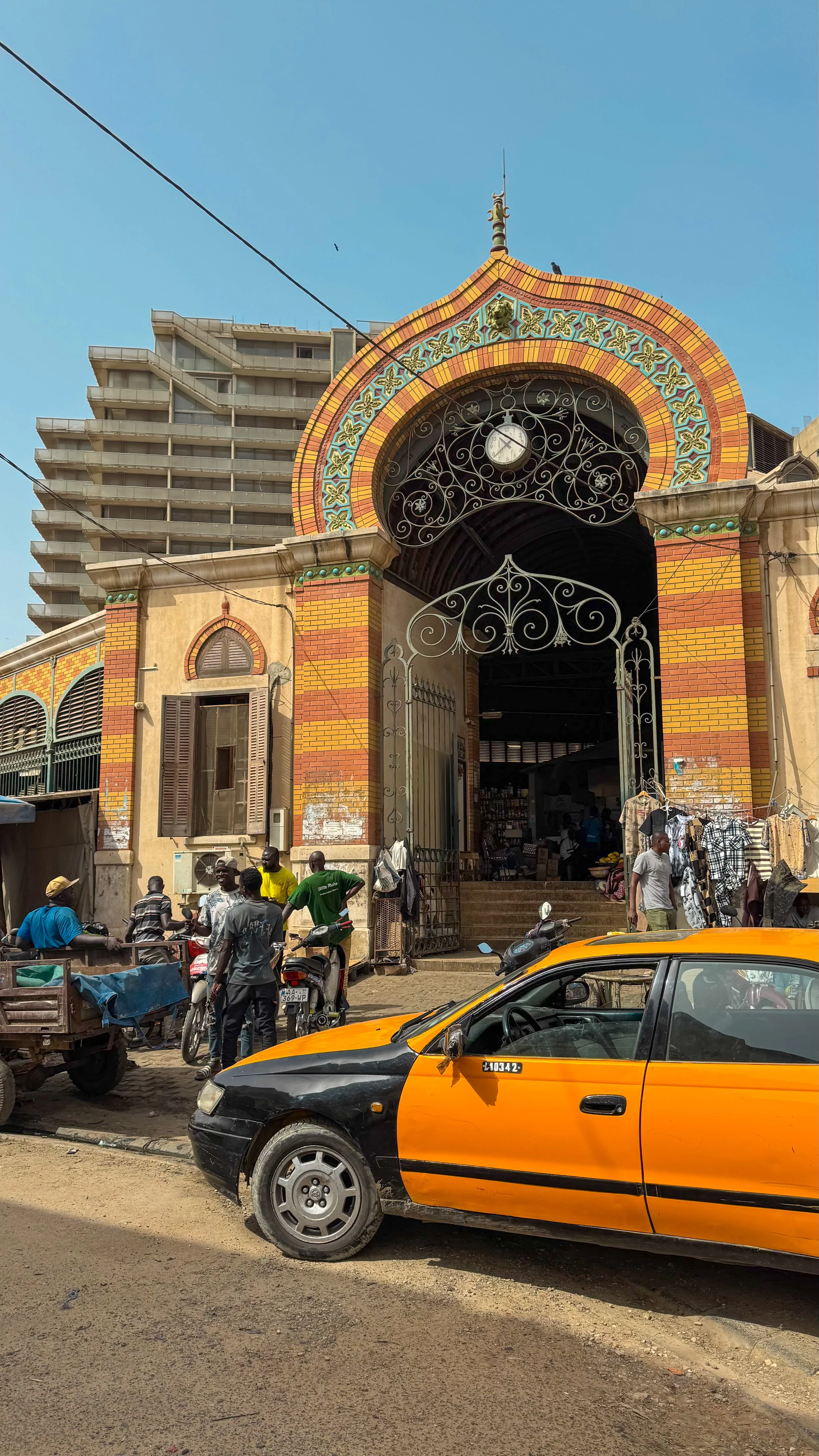 Une scène urbaine avec un bâtiment ancien avec une horloge, un portail en fer forgé et des personnes autour, des motos, une voiture jaune et des stands de vente en plein air.