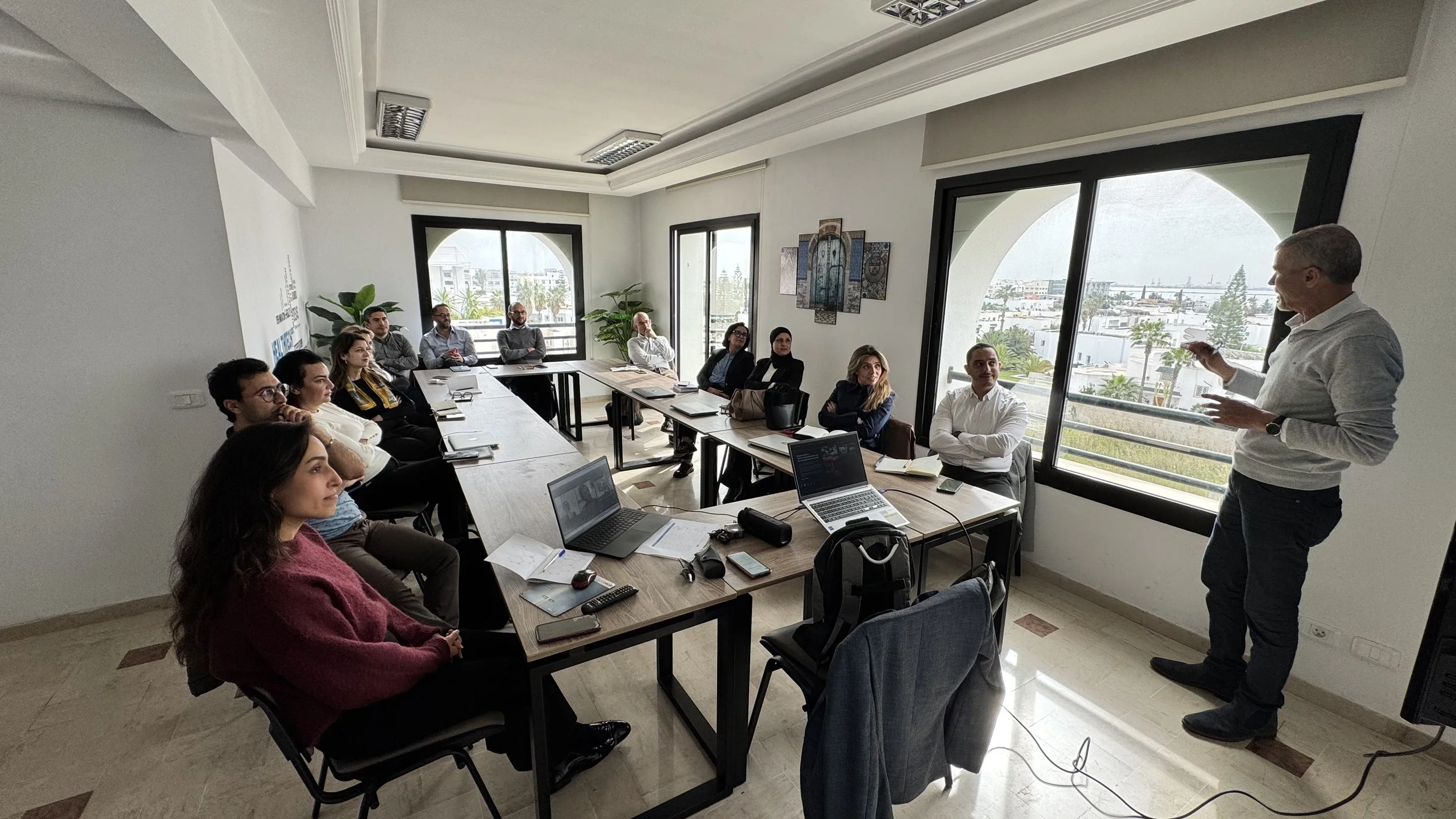 Une réunion dans une salle de conférence avec plusieurs personnes assises autour d'une grande table et un homme debout en train de parler, avec vue sur la ville depuis de grandes fenêtres.