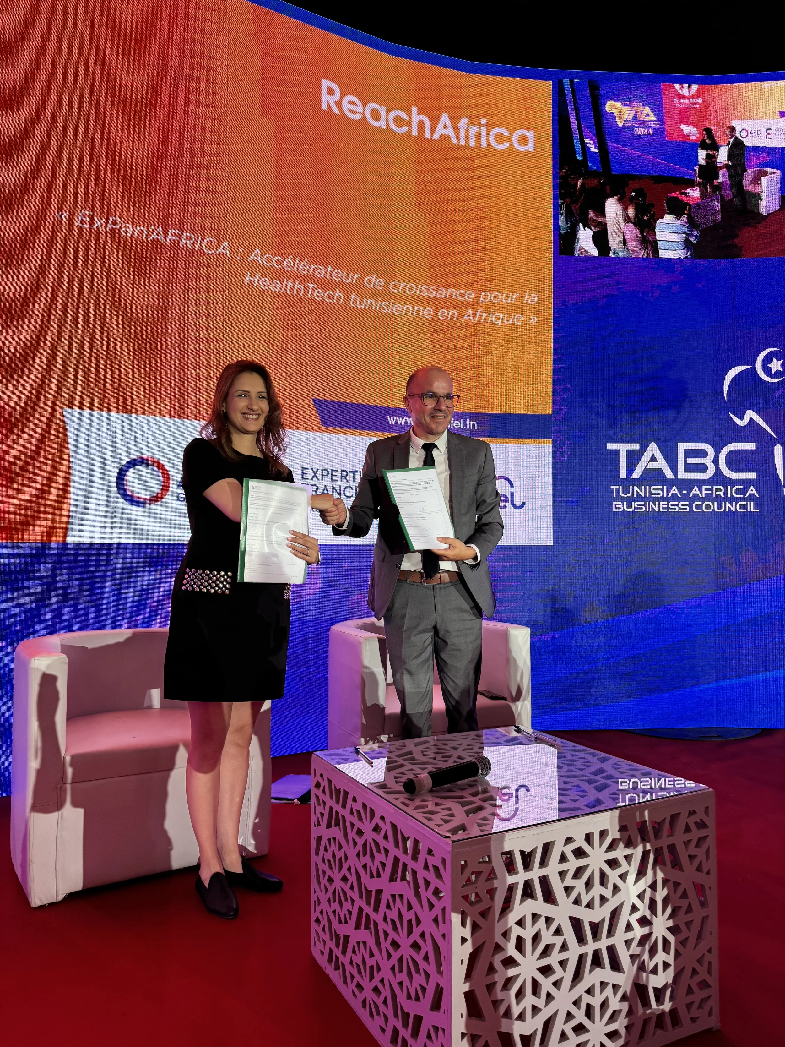 Deux personnes, une femme et un homme, se tiennent sur scène, tenant des documents, lors d'une cérémonie. En arrière-plan, un grand écran affiche le texte 'ReachaAfrica' et des logos liés à la Tunisie et à l'Afrique.