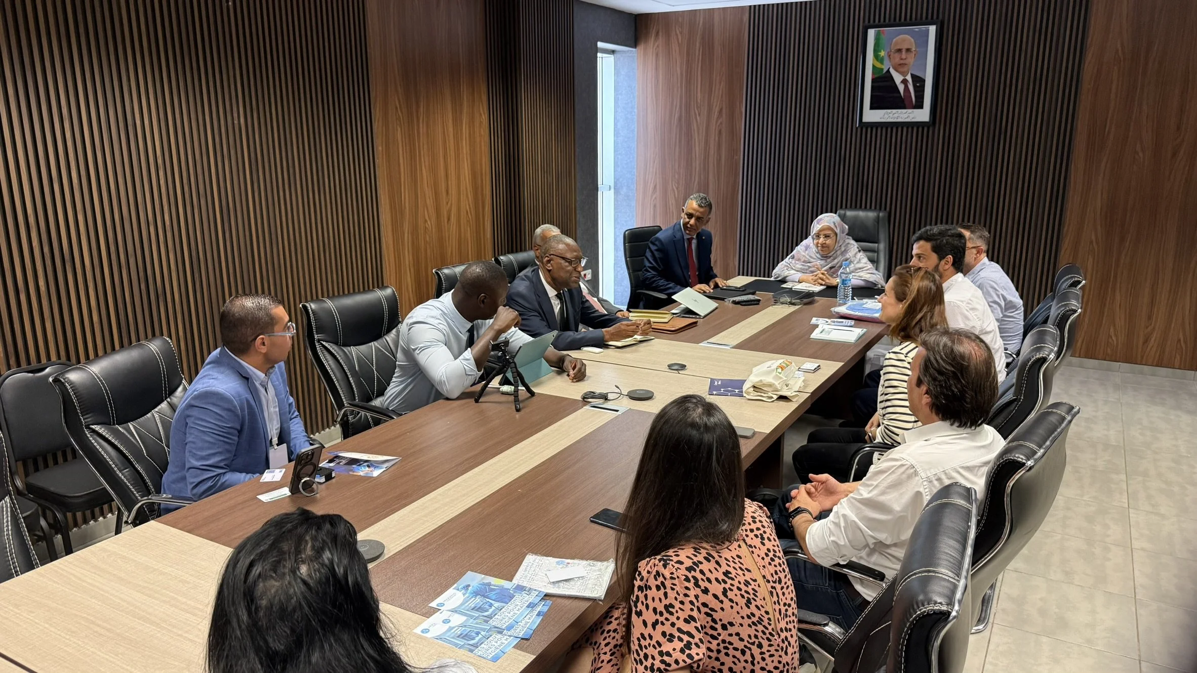 Une réunion formelle avec plusieurs personnes assises autour d'une grande table en bois dans une salle moderne. Parmi eux, on distingue un homme en costume noir, une femme en hijab, et d'autres personnes en tenue professionnelle. Sur le mur, un portrait d'une personne avec drapeau en arrière-plan.