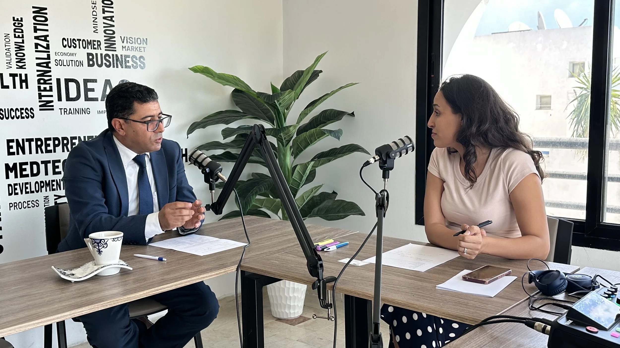Deux personnes, un homme en costume et une femme en blouse, discutent lors d'enregistrement dans une salle de podcast ou interview, avec microphones et matériel audio, en intérieur près d'une fenêtre avec vue sur un immeuble et un arbre, décorée de mots liés aux affaires et à l'innovation sur un mur.
