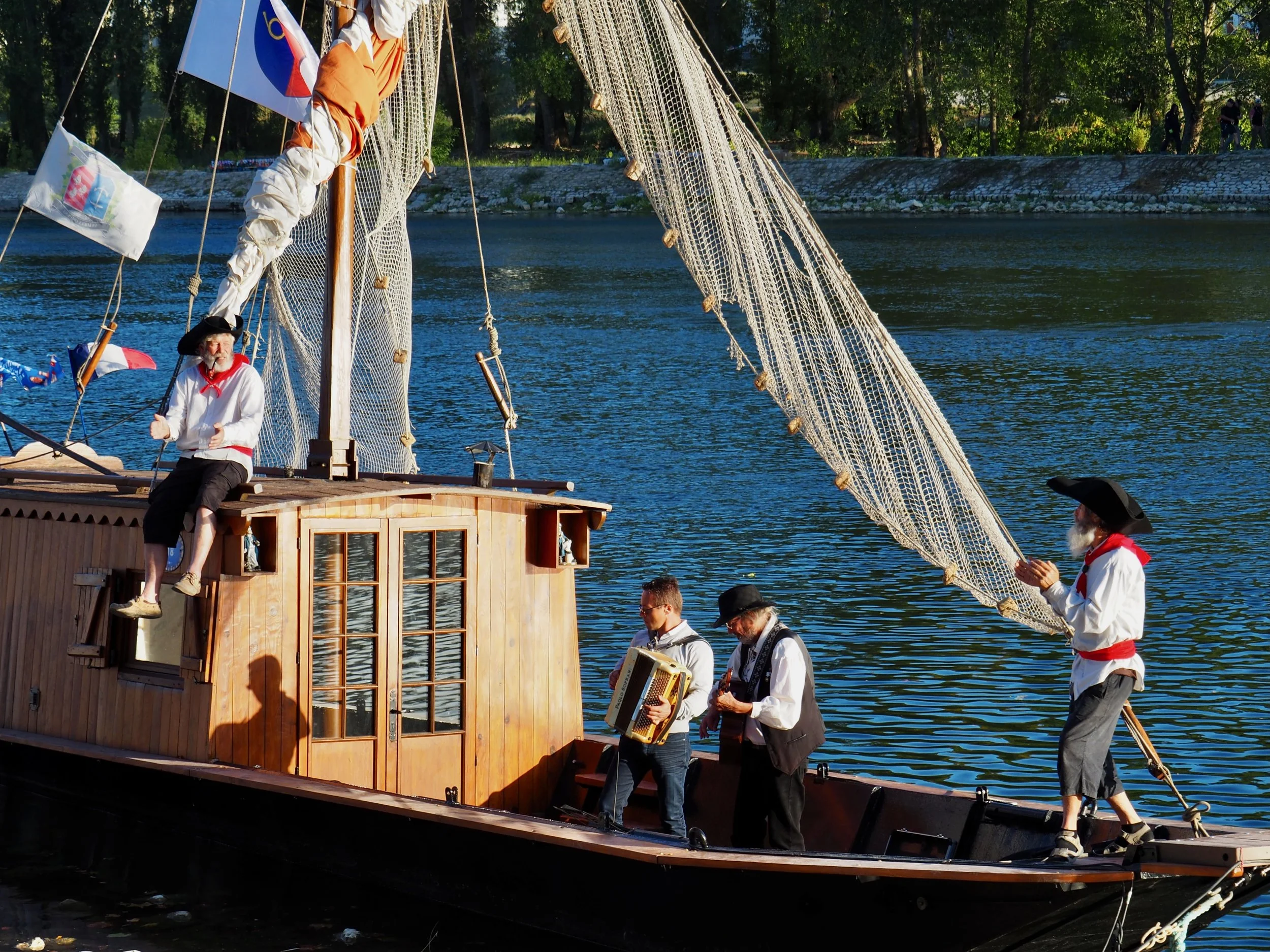 Une scène de théâtre de rue sur un bateau en bois avec des personnes habillées en costumes historiques, jouant des instruments, sous un soleil brillant avec des arbres en arrière-plan.