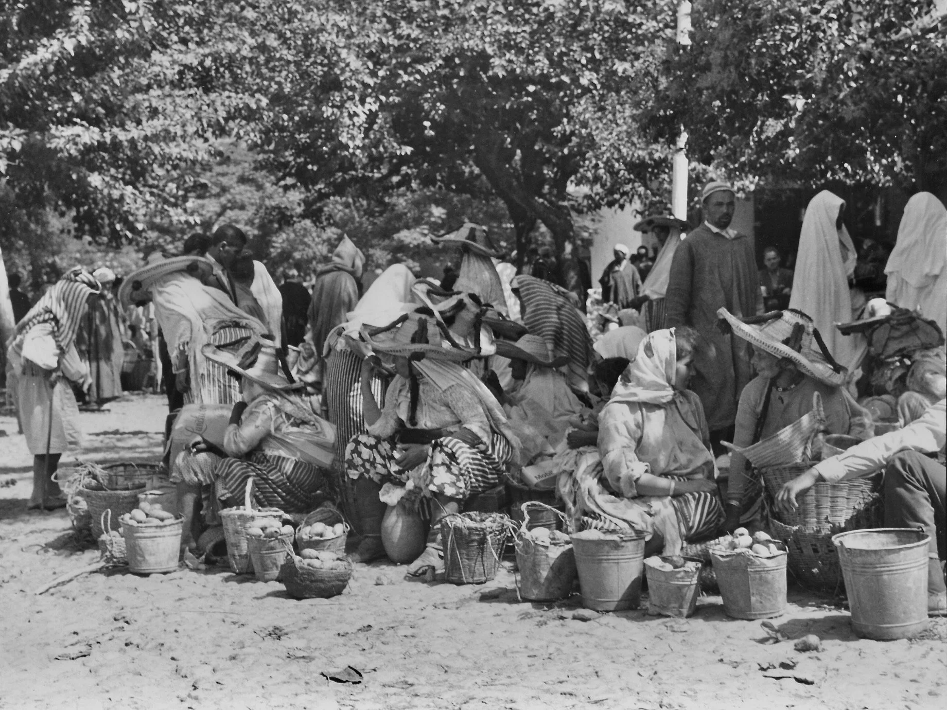 Marché en plein air avec des femmes portant des vêtements traditionnels, paniers remplis de fruits et légumes, sous des arbres.
