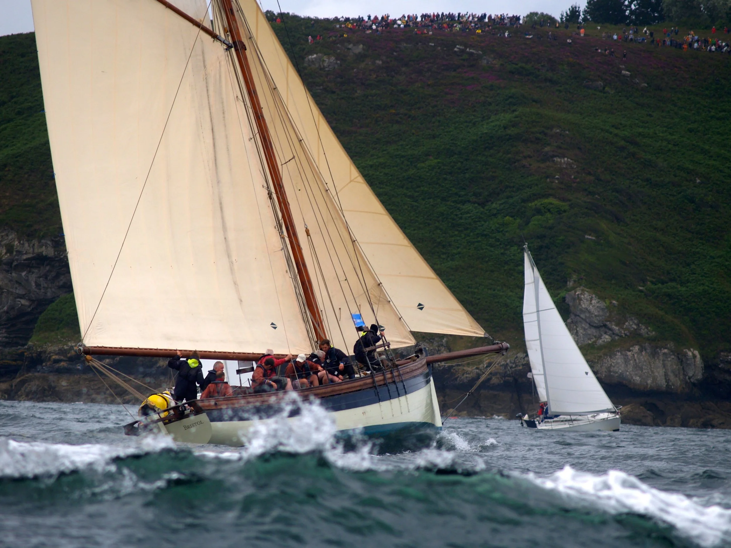 Un voilier avec plusieurs personnes à bord naviguant dans l'eau, avec une colline verdoyante et une foule de spectateurs au sommet en arrière-plan.