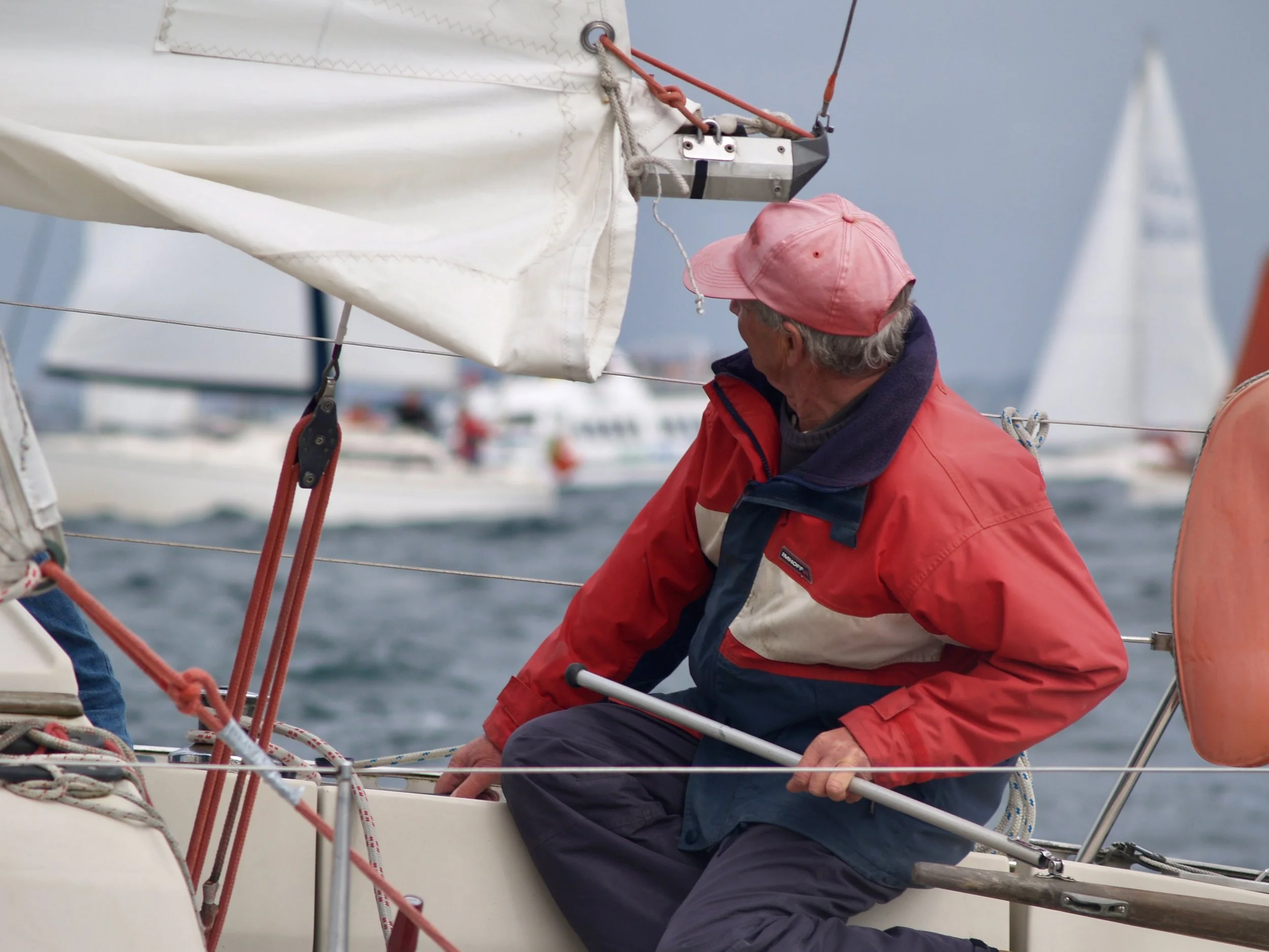Un homme portant une casquette rose et une veste rouge travaille sur un voilier, ajustant des cordages et une voile.