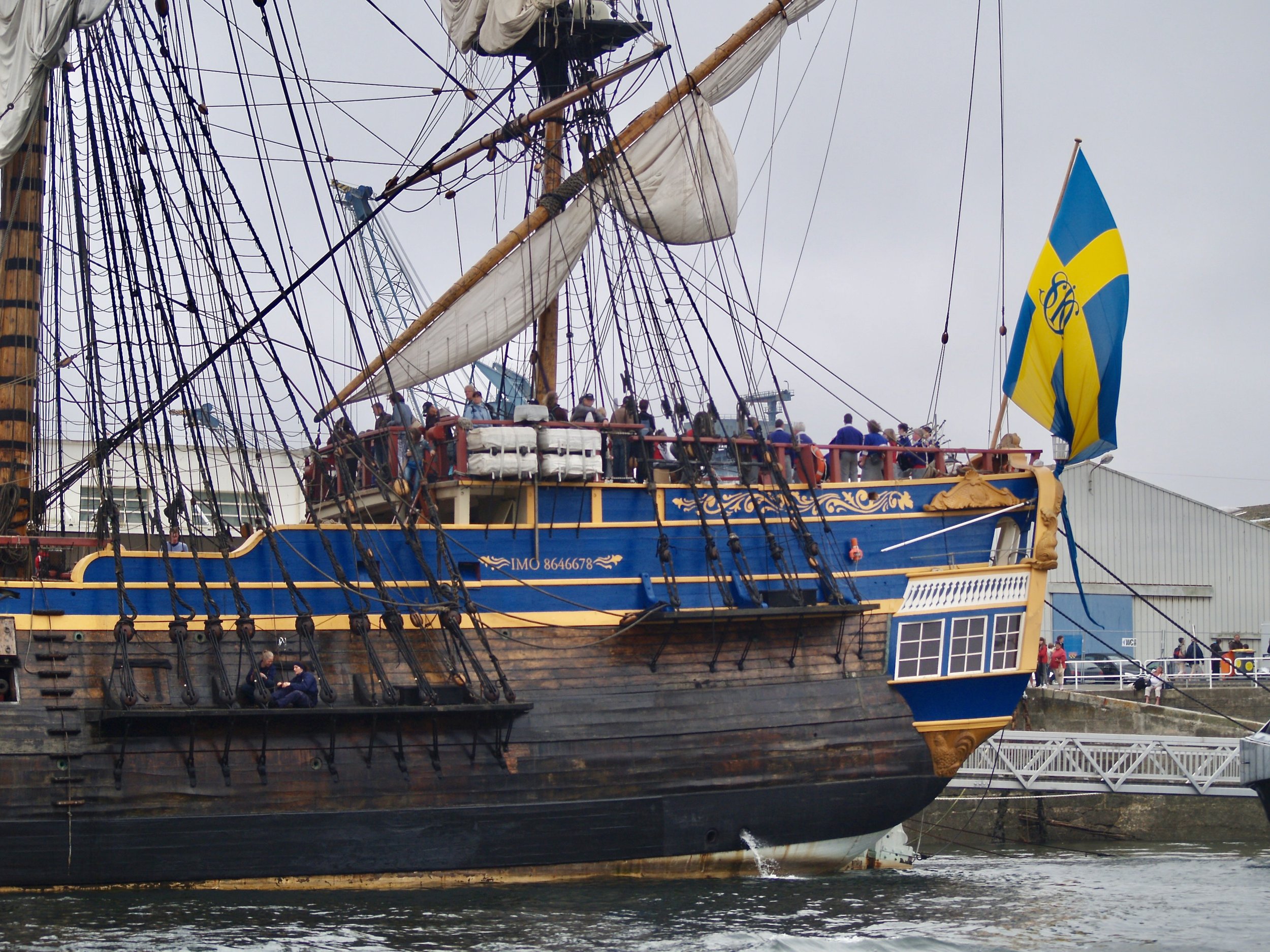 Un grand navire ancien en bois avec des cordages noirs complexes, une voile partiellement déployée, et un drapeau suédois flottant. Des personnes sont visibles sur le pont. Le navire est amarré au port.