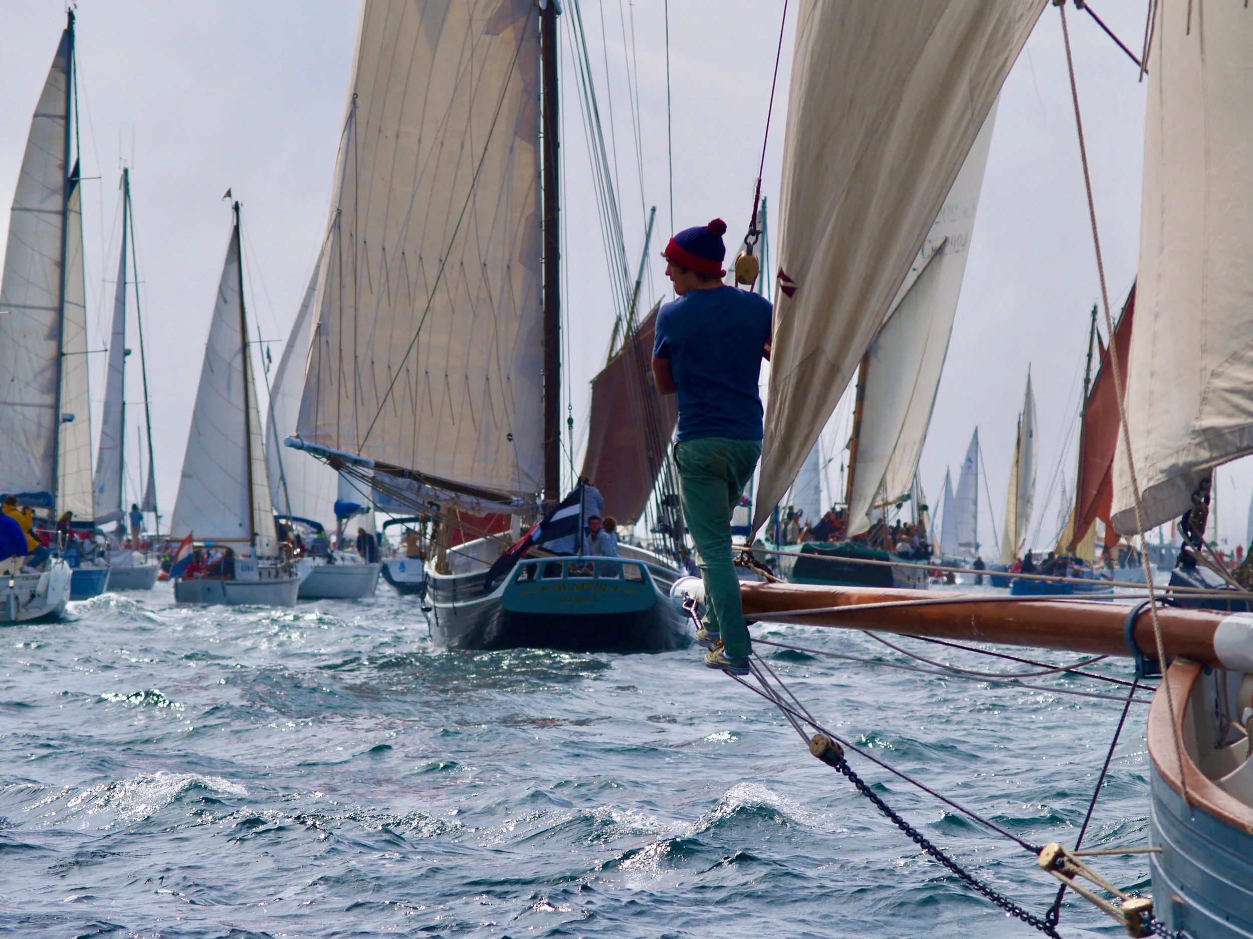 Jeune homme en t-shirt bleu et bonnet à pompon rouge et bleu, debout sur un bateau à voile, parmi une flotte de voiliers en mer sous un ciel nuageux.