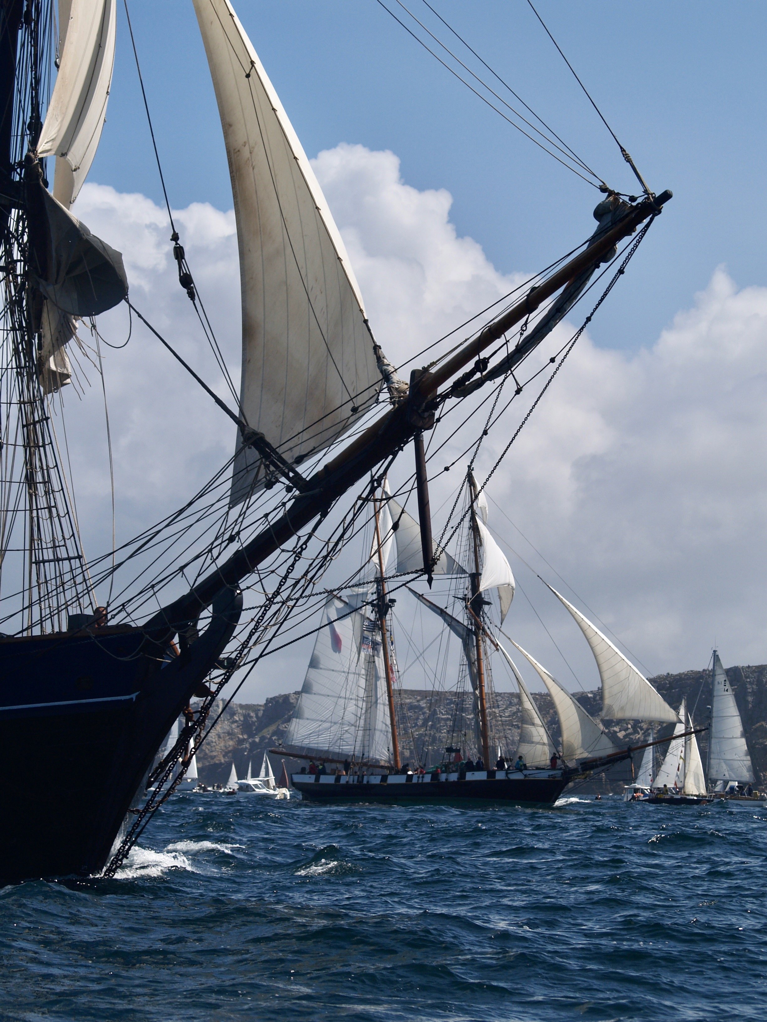 Bateaux à voile naviguant en mer avec des voiles blanches déployées, sous un ciel partiellement nuageux.
